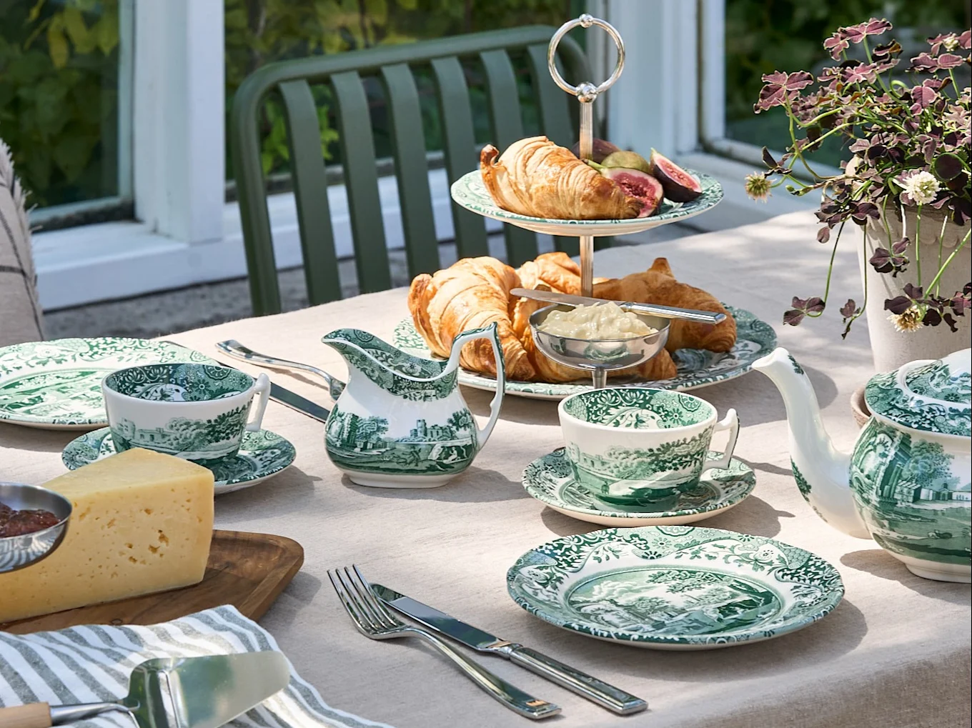 Een buitentafel gedekt met groen-wit servies, croissants, kaas en een theepot, in een tuinomgeving.