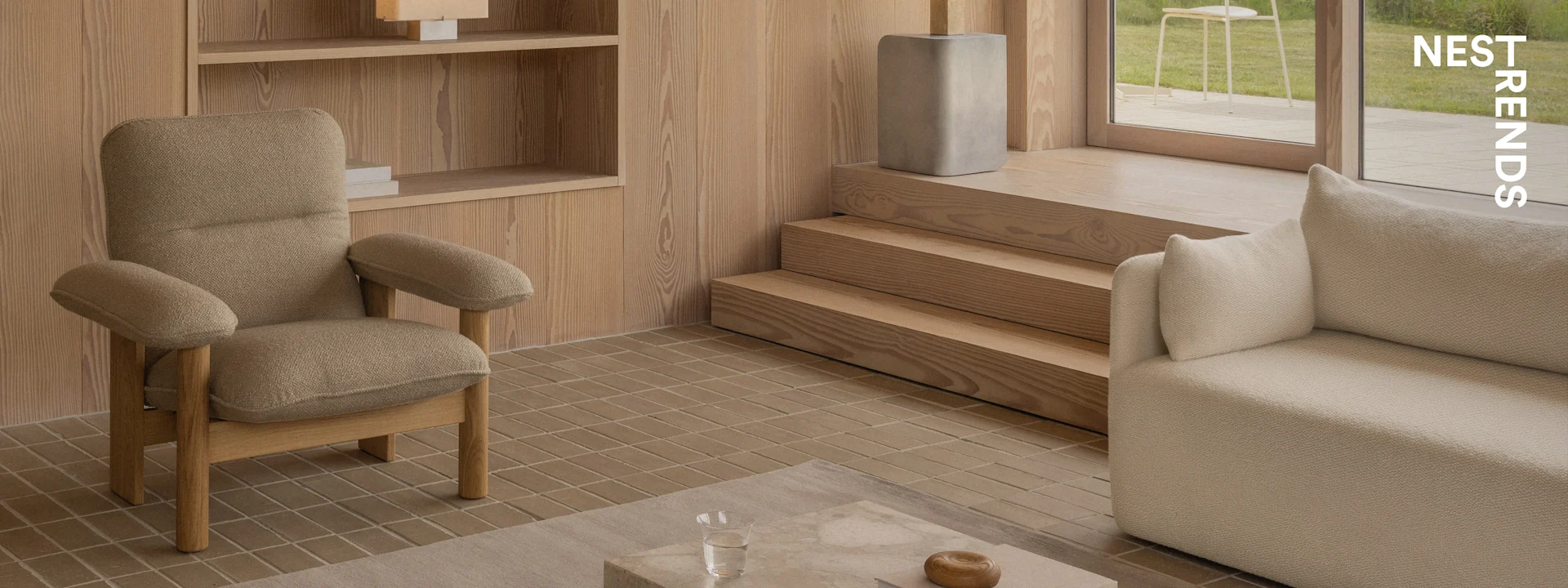 Een moderne woonkamer met een getextureerde bruine fauteuil, een beige bank, houten wandpanelen, een tegelvloer en uitzicht op een tuin.
