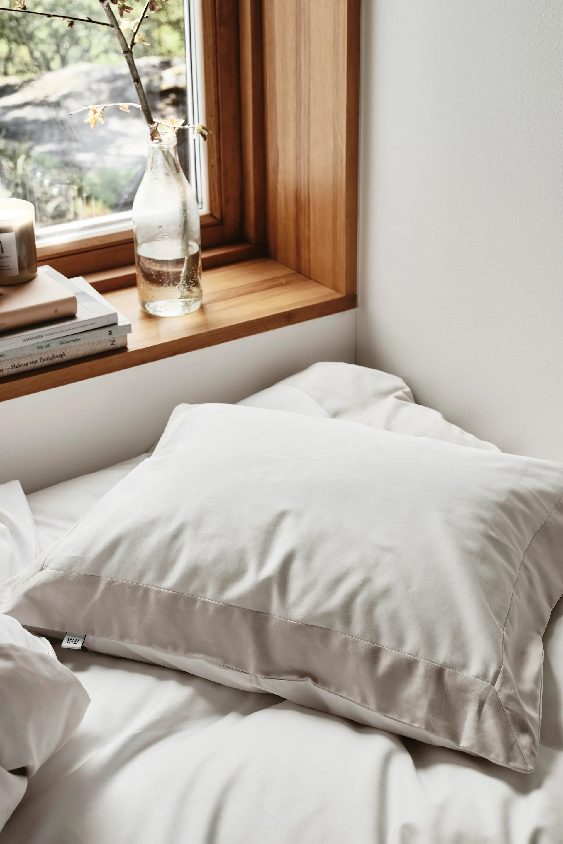 Een lichtbeige kussen op een wit bed, naast een raam met een houten vensterbank, boeken en een plantentak in een fles.