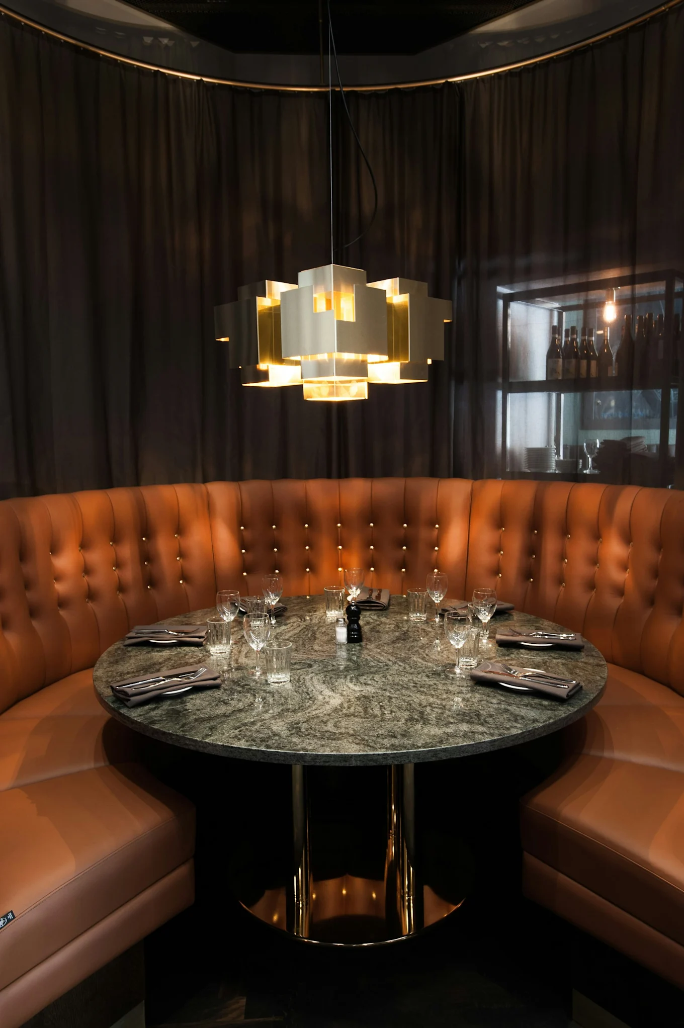 Een privé-eethoek met een gebogen bruine lederen bank, een ronde marmeren tafel en een moderne gouden geometrische kroonluchter.