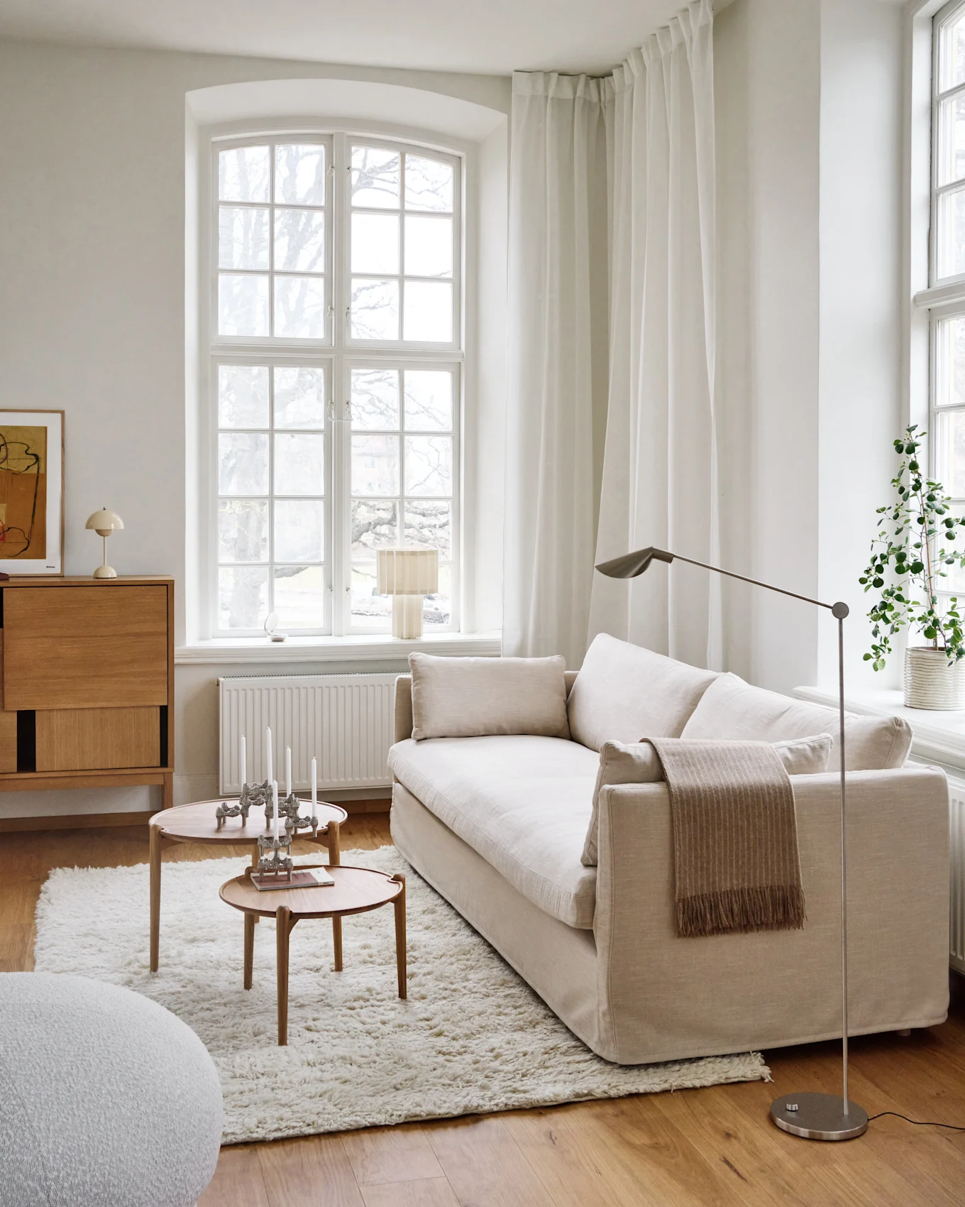 Een lichte, minimalistische woonkamer met een lichtbeige bank, twee ronde houten salontafels met kaarsen, een shaggy wit vloerkleed en een groot raam.