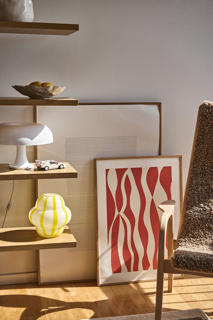 De Curlie-vaas van Byon heeft een ronde, bolvormige ontwerp in geel- en wit gestreept glas. Hier staat deze in een open boekenplank, samen met een Shell-schaal, lampen en schilderijen.