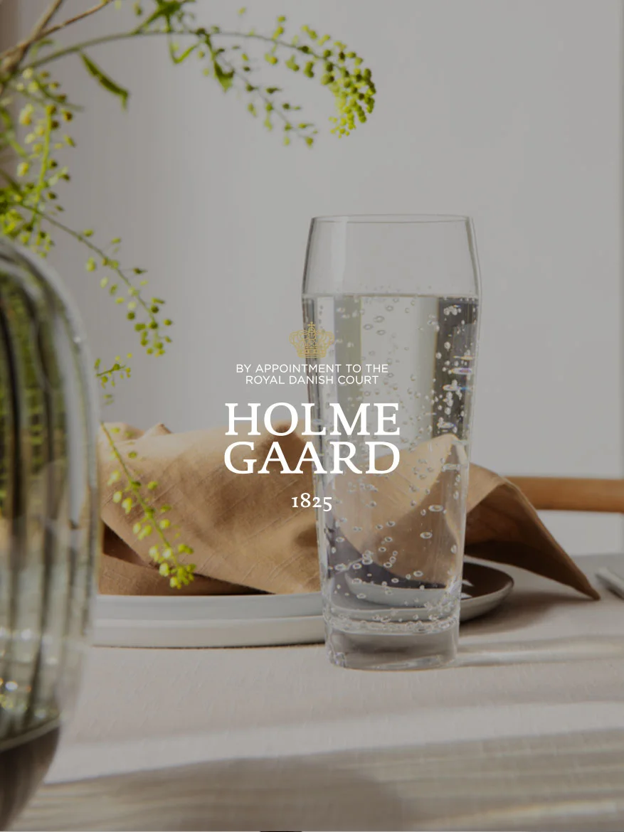 Een hoog glas bruisend water naast een bruine servet en een plant, met het Holmegaard-logo eroverheen.