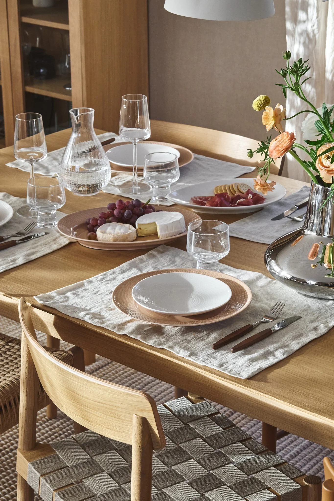 Houten eettafel met borden, glazen, waterkan, druiven, kazen, vleeswaren en bloemen. Gevlochten stoelen.