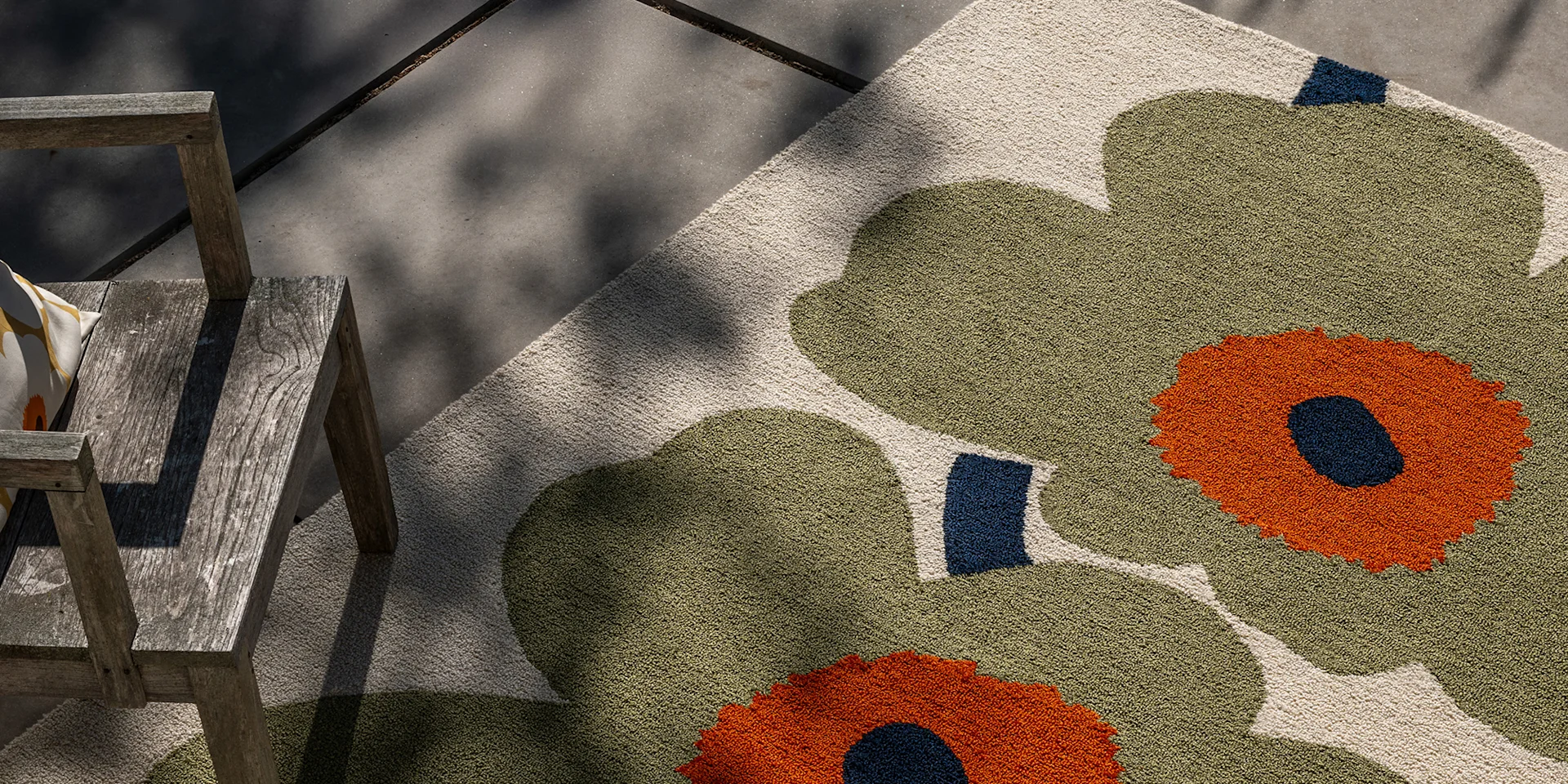 Olijfgroen en oranje bloemen buitenkleed naast een verweerde houten stoel op een betonnen terras.