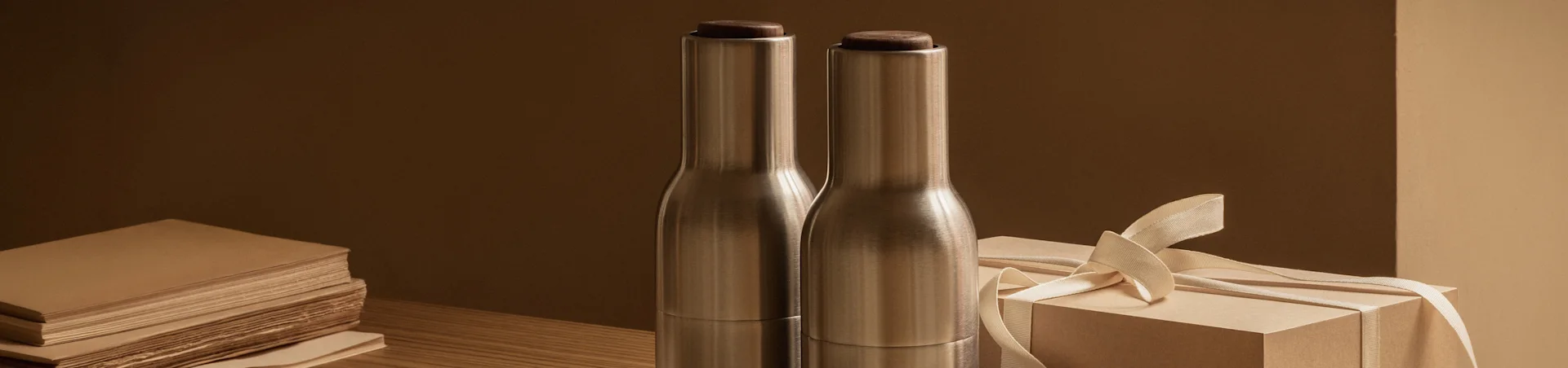 Twee geborstelde metalen flessen met donkere houten doppen naast een lichtbruine geschenkdoos met lint op een houten tafel.