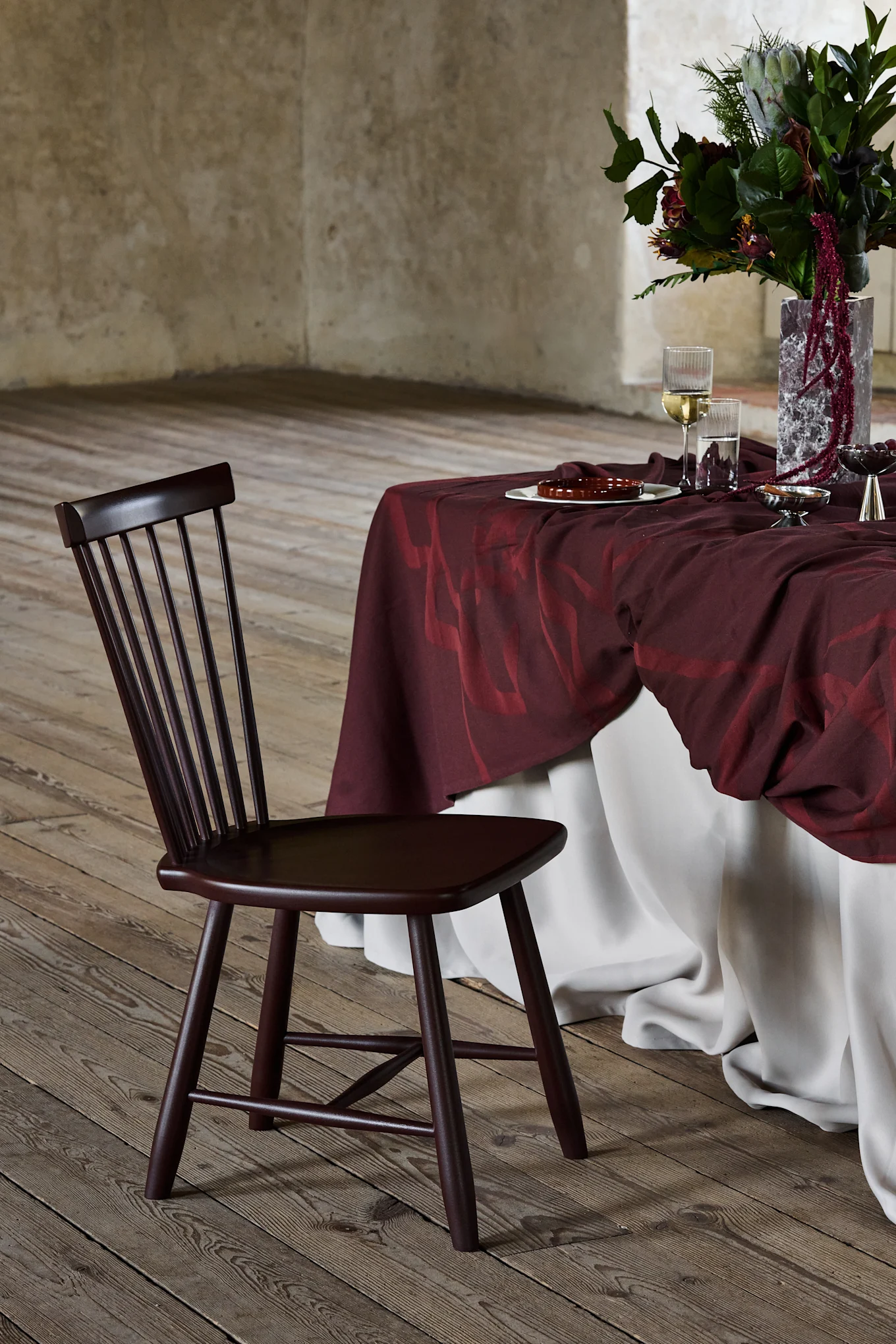 Donkerbruine houten stoel met spijlenrug, naast een tafel met een bordeauxrood tafelkleed en bloemstuk, op een houten vloer.