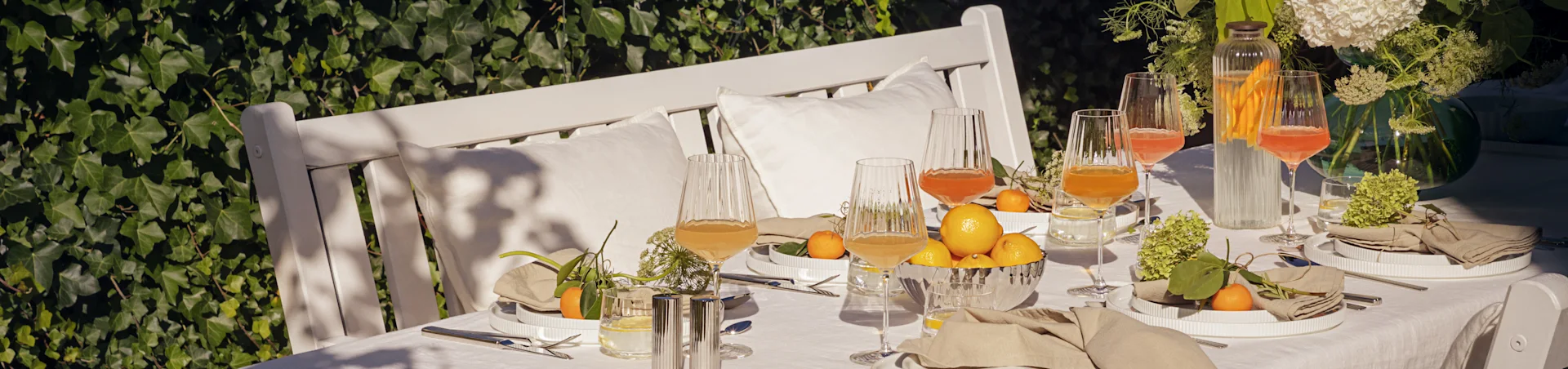 Buitentafel gedekt voor zomermaaltijd met witte geribbelde borden, linnen servetten, geribbelde glazen, citroenen en mandarijnen.