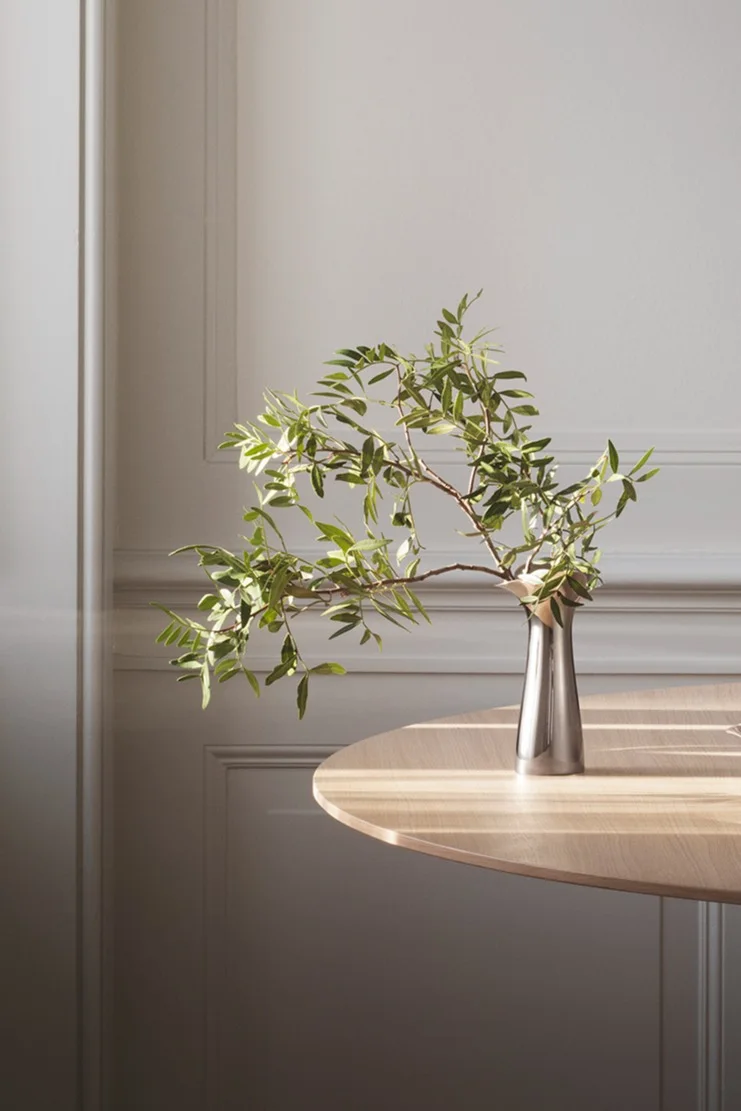 Zilveren vaas met groene bladrijke takken op een lichte houten tafel, zonlicht werpt schaduwen op tafel en muur met sierlijsten.