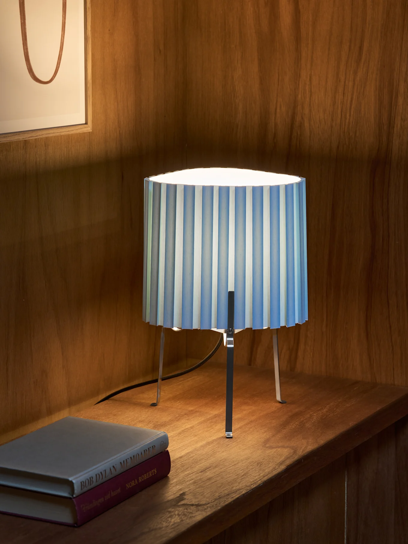 Blauw-wit gestreepte geplooide tafellamp met drie metalen poten op houten plank naast twee boeken. Ingelijst kunstwerk op achtergrond.