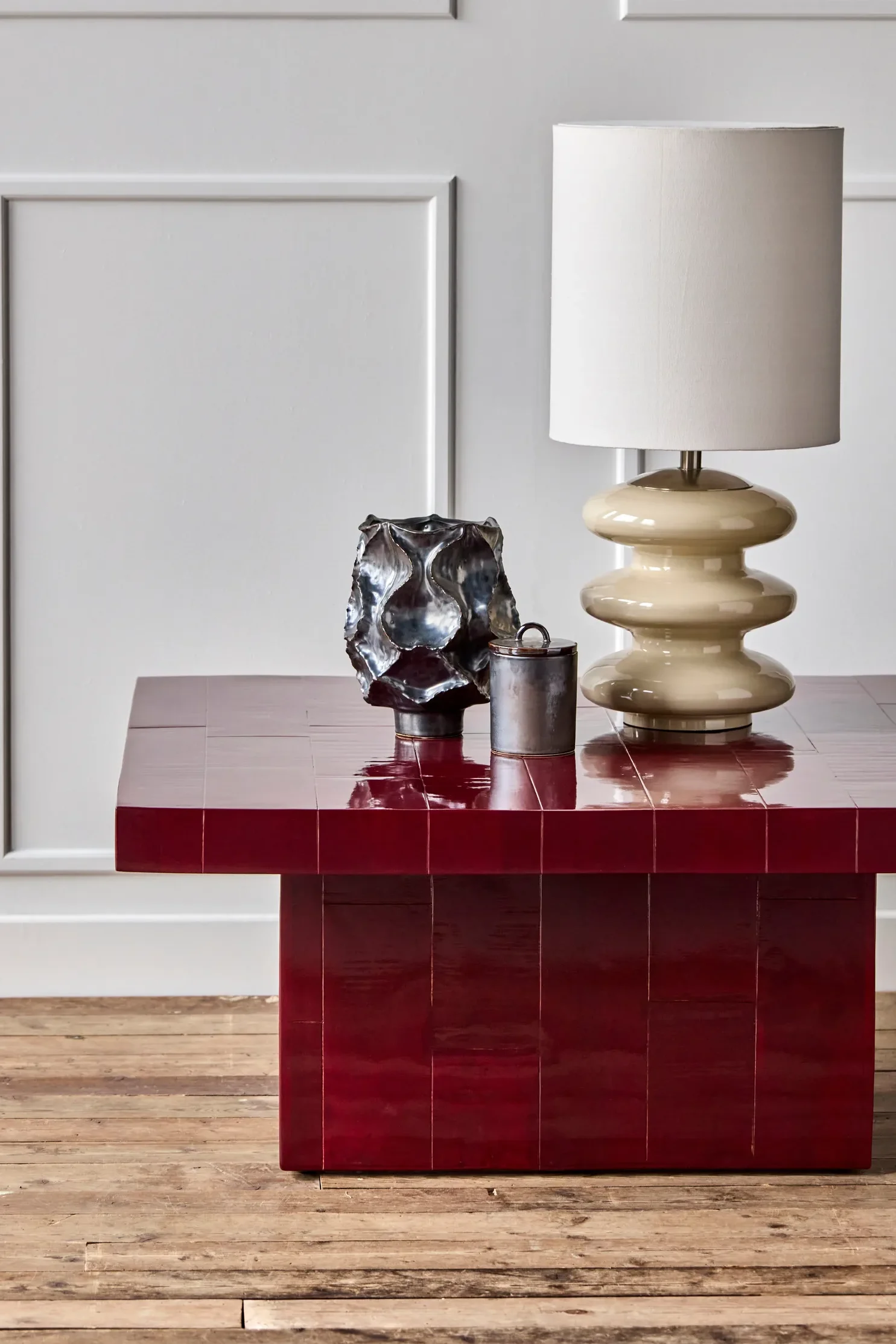 Glanzende donkerrode tegel salontafel met een crèmekleurige sculpturale lamp, een donkere metalen abstracte vaas en een kleine cilindrische houder.