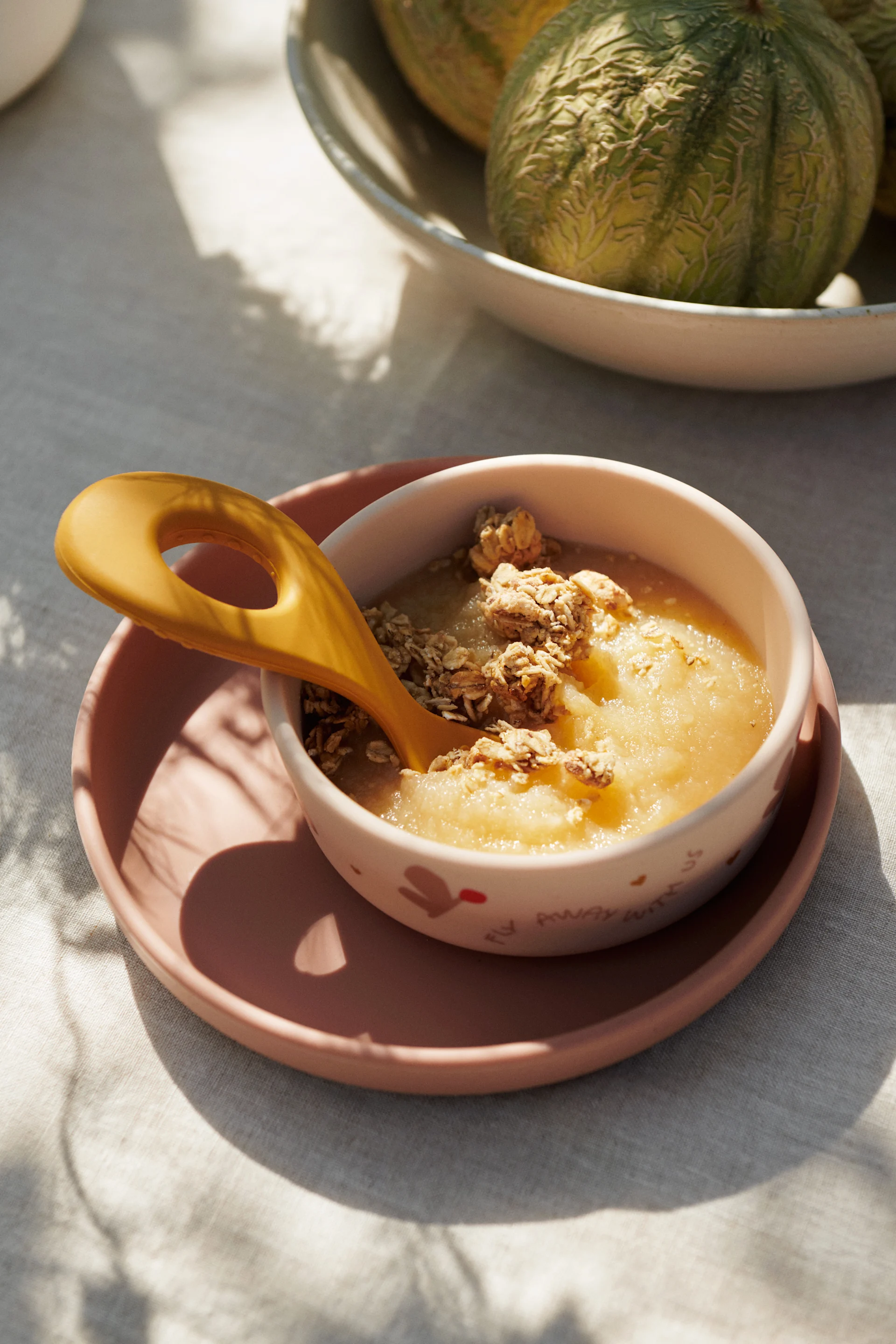 Appelmoes met granola en een gele lepel in een kom op een roze bord, met meloenen.