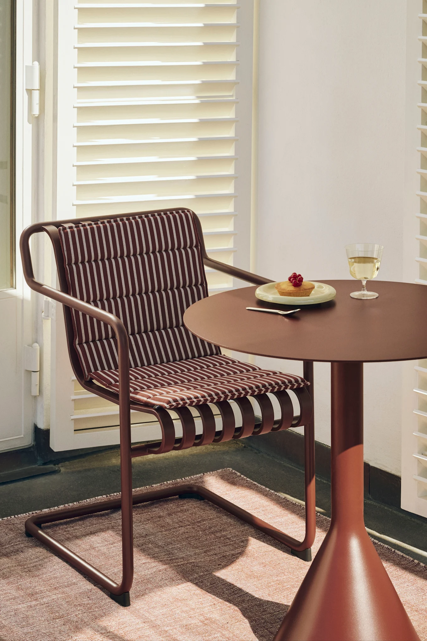 Een roodbruine stoel met gestreept kussen naast een bijpassende ronde tafel met een taartje en een glas witte wijn.