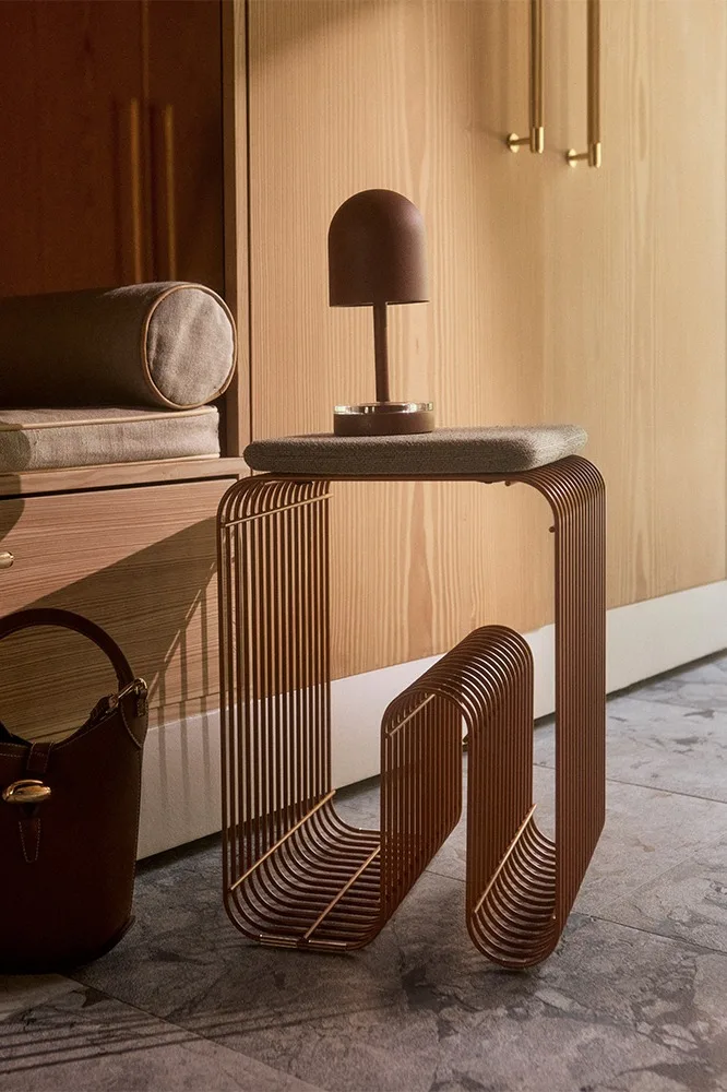 Moderne bijzettafel van bruine verticale metalen staven met een bruine koepellamp en handtas, tegen een houten kast.