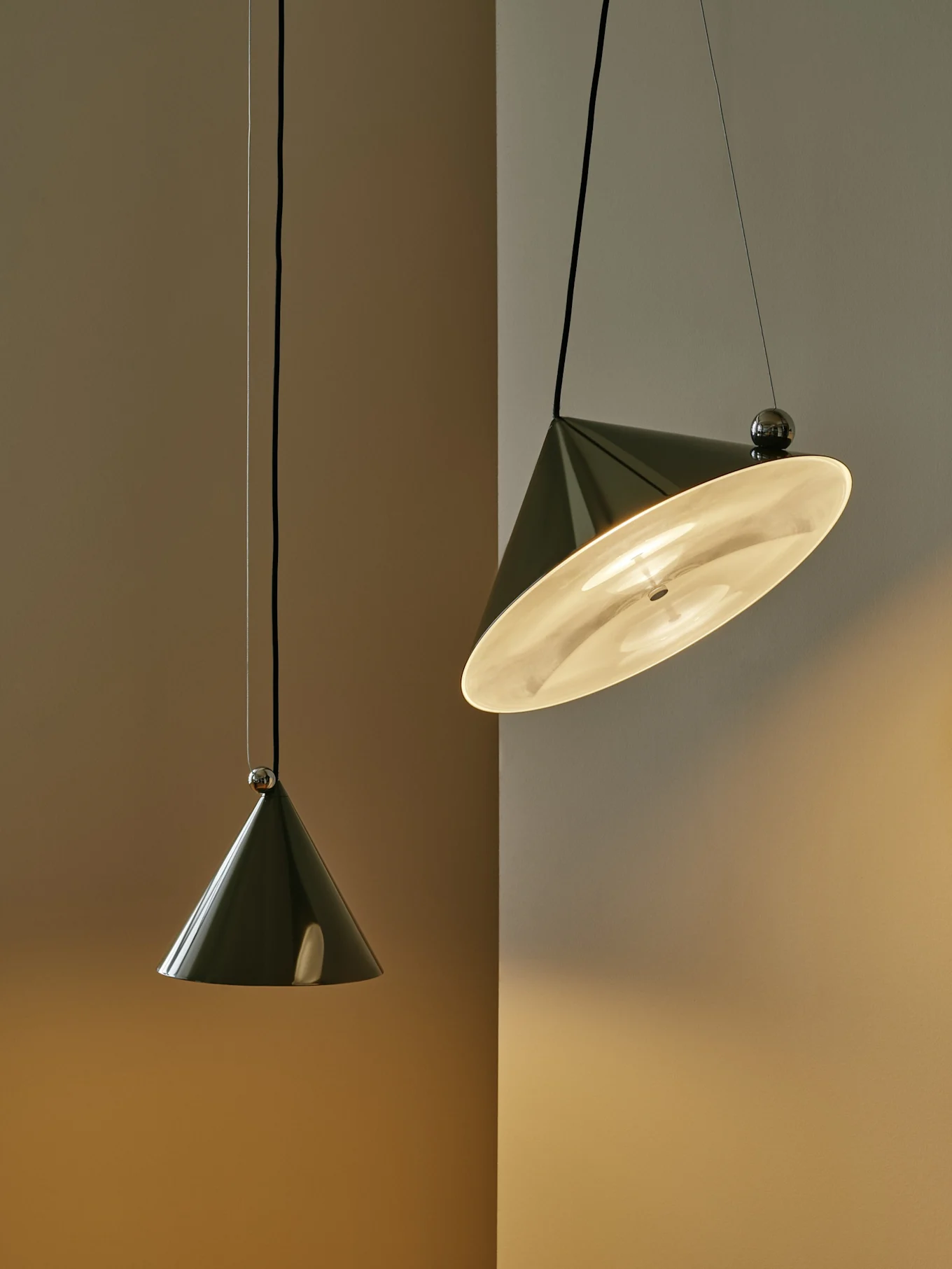 Twee donkergroene conische hanglampen tegen een tweekleurige muur; één is verlicht en gekanteld, de andere hangt verticaal.