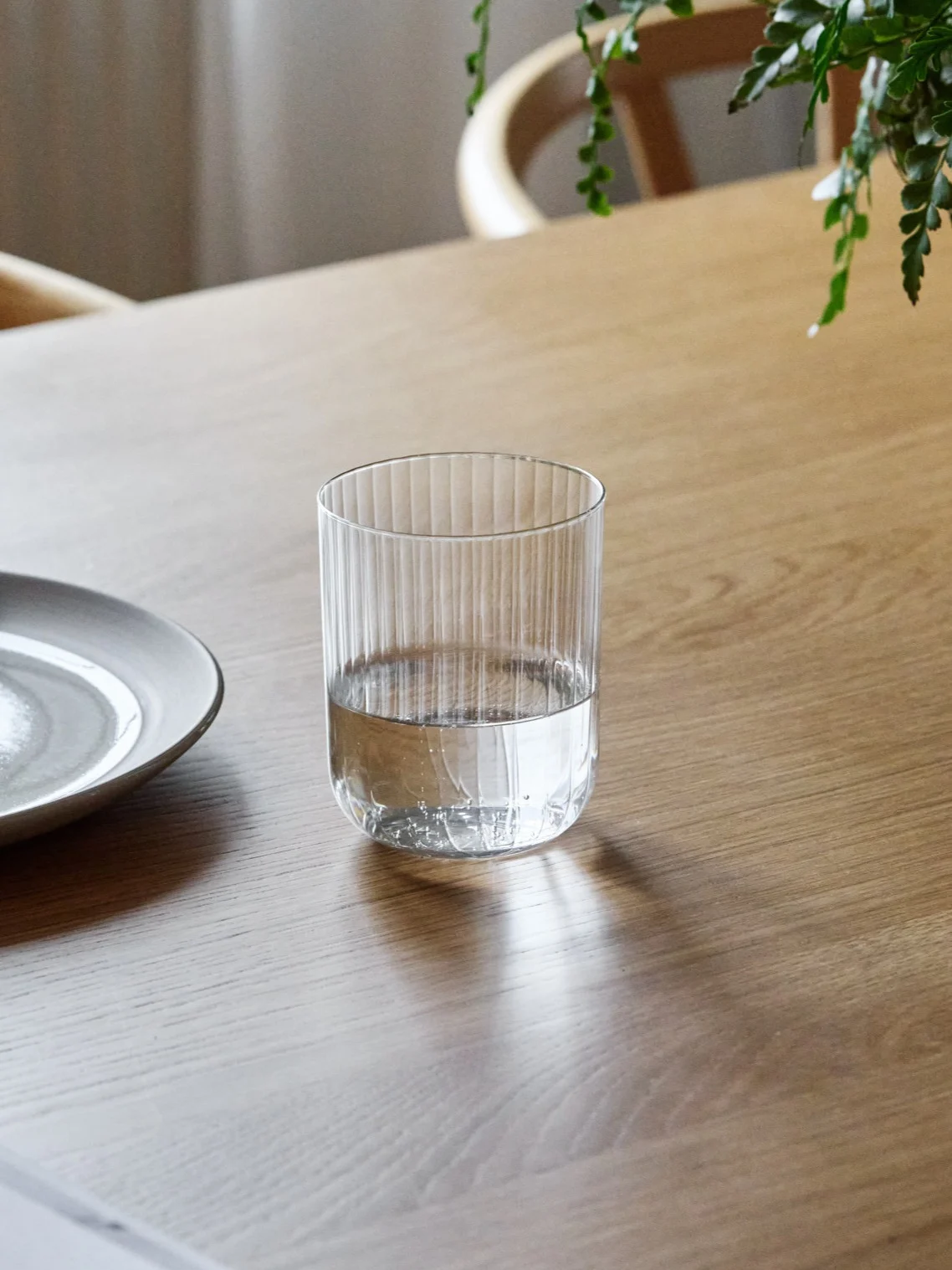 Een geribbeld glas, halfvol met water, staat op een houten tafel naast een keramisch bord.
