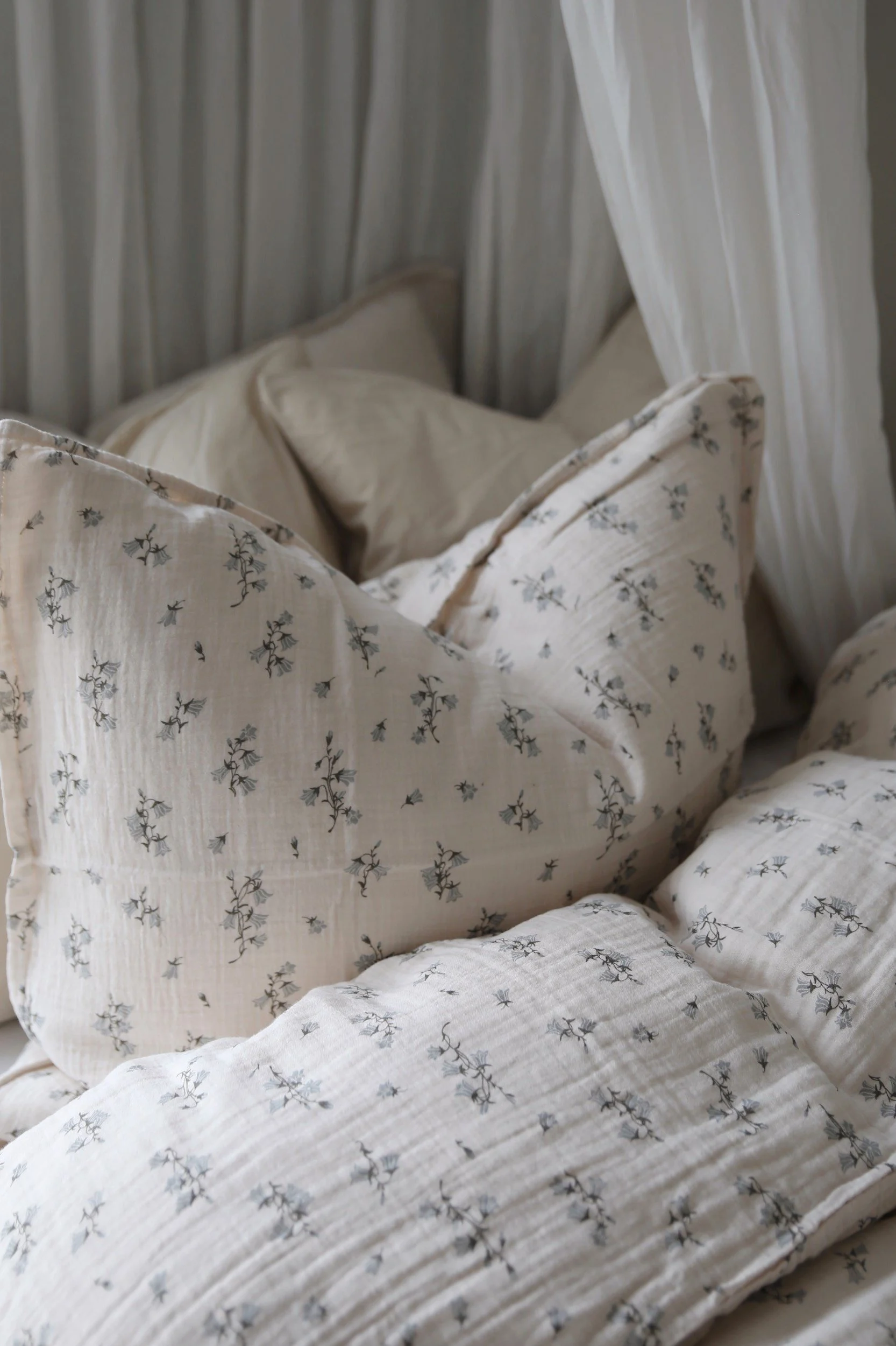 Gezellig beddengoed van lichtbeige kreukelstof met een delicaat blauw bloemendessin en witte gordijnen.