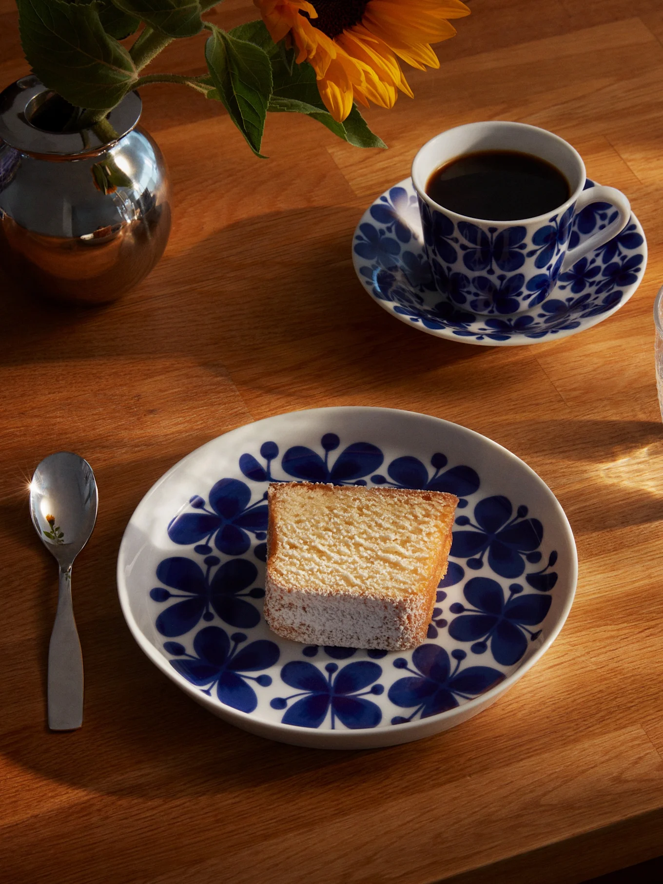 Een stuk cake op een blauw-wit bloemenbord, met een koffiekop, een glas water, een lepel en een zonnebloem in een zilveren vaas op een zonnige houten tafel.