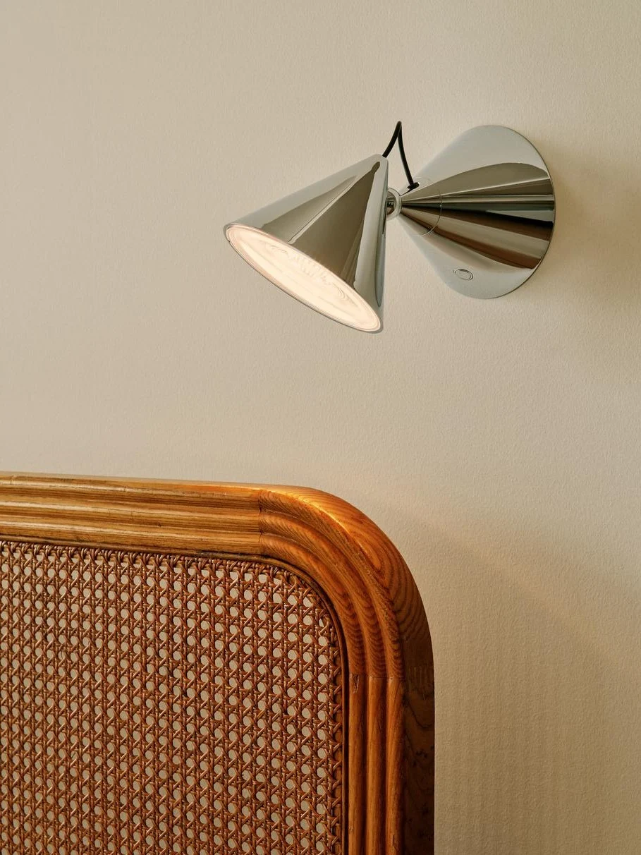 Moderne chromen wandlamp met conische kap verlicht een lichte muur boven een geweven rotan en houten hoofdeinde.