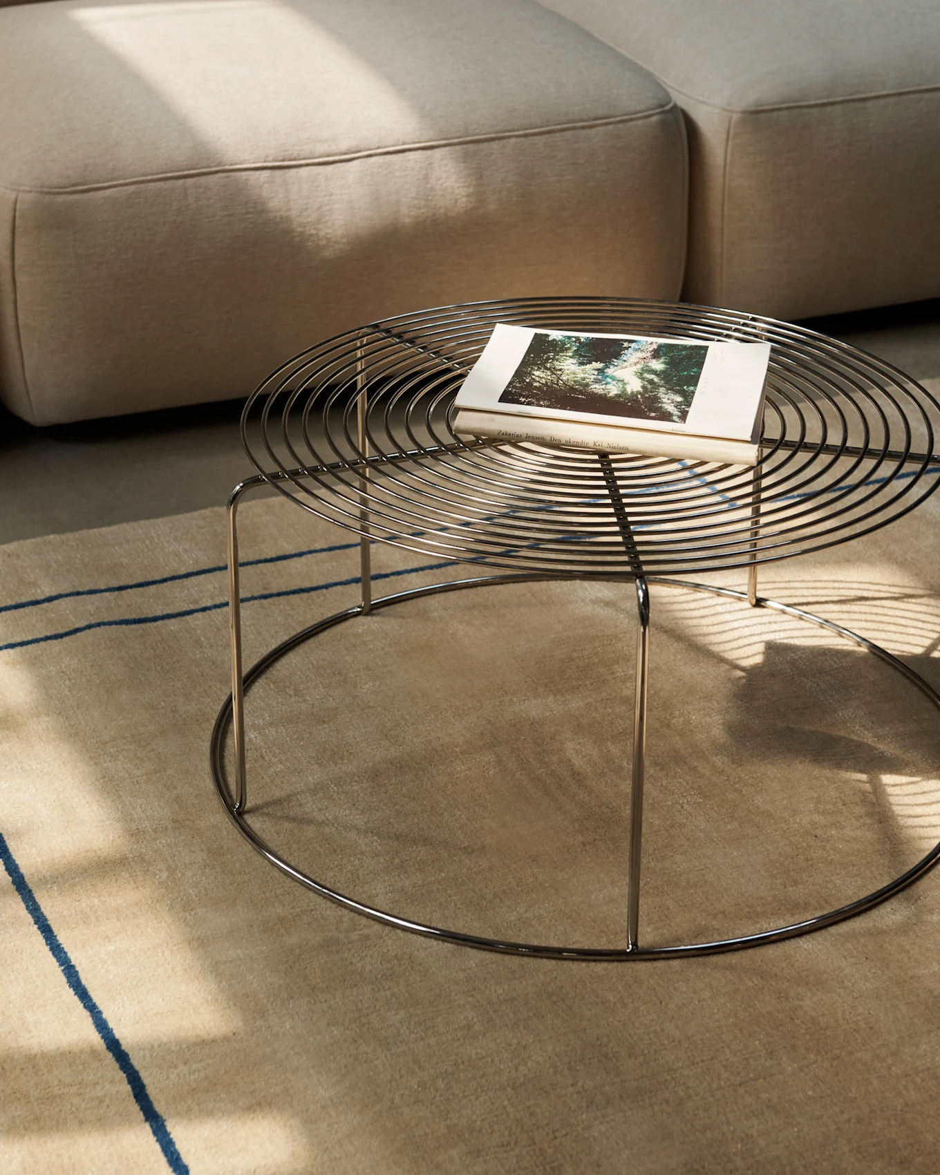 Ronde metalen salontafel met boek op een beige tapijt, naast een bank.