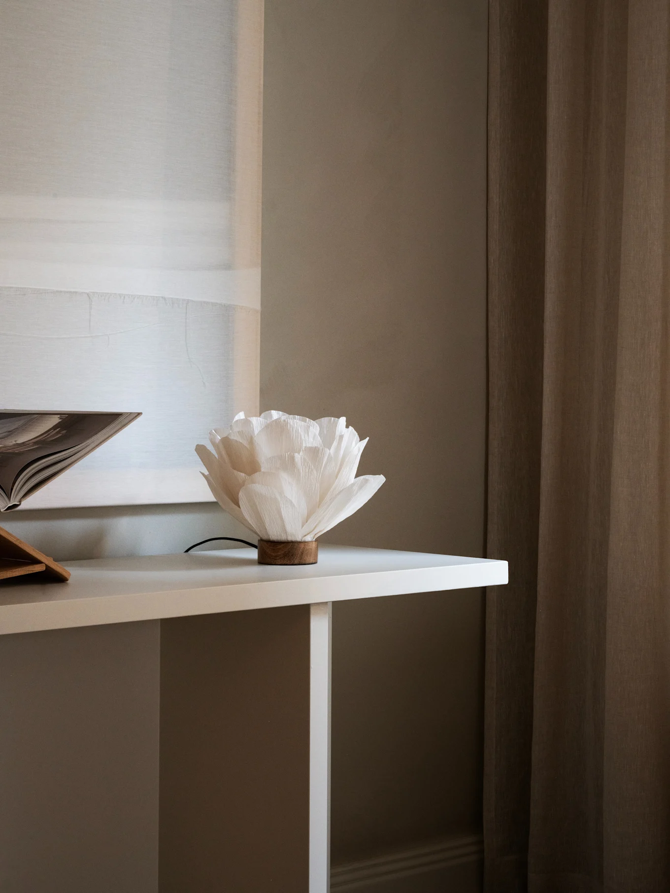 Witte bloemvormige papieren lamp met een houten voet op een witte tafel, naast een open boek. Bruine gordijnen op de achtergrond.