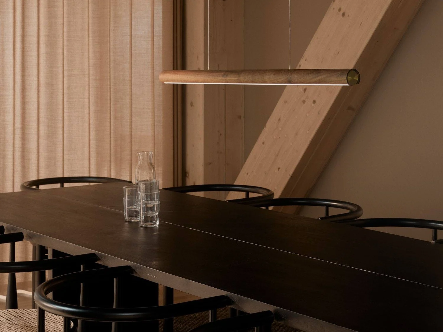 Een moderne eetkamer met een lange donkere houten tafel, zwarte stoelen en een slanke houten hanglamp. Een glazen karaf en drie glazen staan op de tafel.