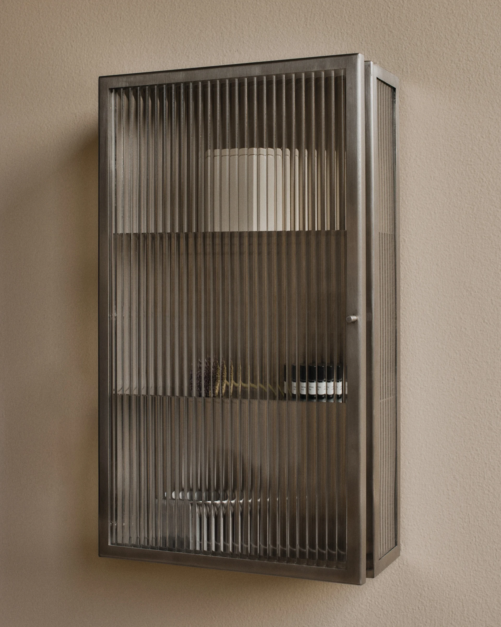 Wandgemonteerde metalen kast met geribbelde glazen deuren, met decoratieve voorwerpen en kleine flesjes.