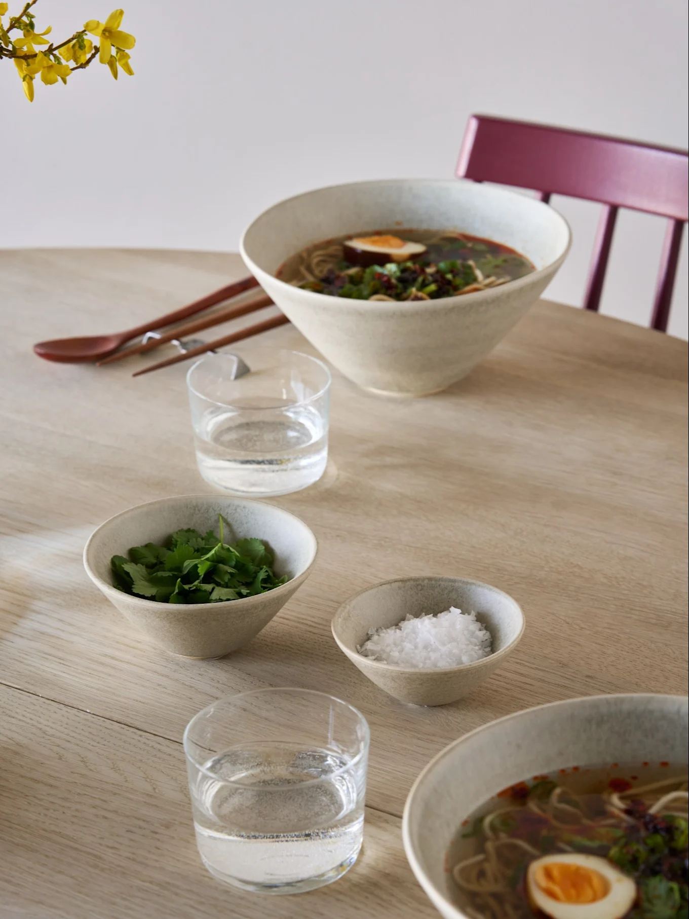 Twee kommen ramen met ei en kruiden, bijgerechtkommen met koriander en zout, water, eetstokjes en een lepel op een lichte houten tafel.