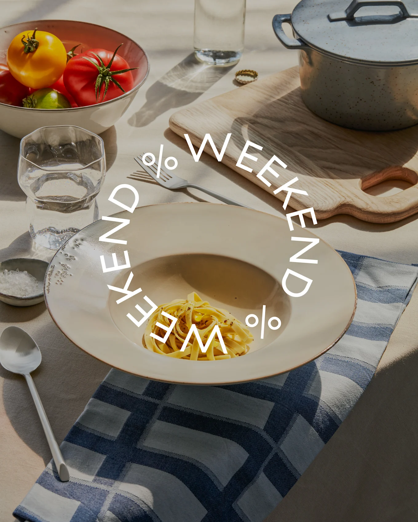 Tafel met pasta, een kom verse tomaten, waterglas en kookpot. De tekst 'WEEKEND % WEEKEND %' is eroverheen geplaatst.