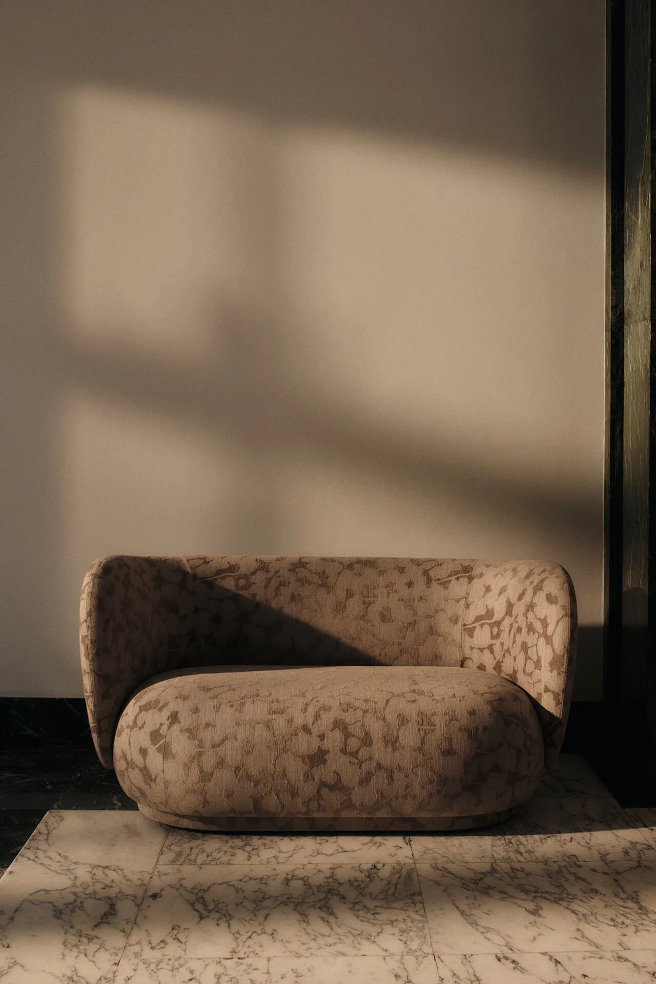 Afgeronde beige fauteuil met subtiel patroon op een witte marmeren vloer, met dramatische schaduwen van warm licht.