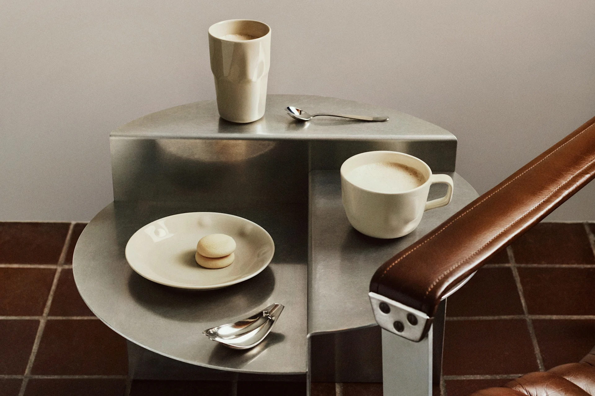 Twee lichtgekleurde koffiemokken, een macaron en lepels op een gelaagde metalen bijzettafel naast een bruine leren fauteuil.