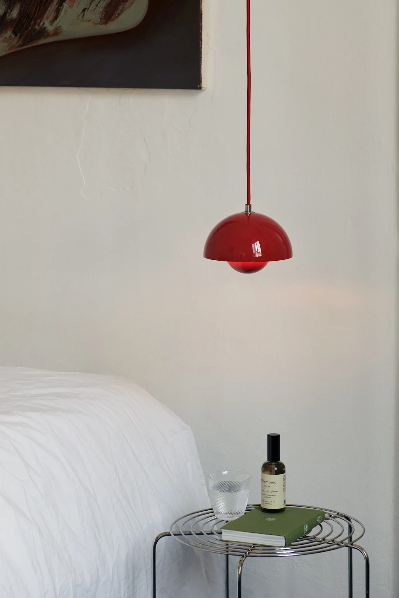 Rode hanglamp boven een chromen draadzijtafel met een glas water, een groen boek en een spuitfles in een slaapkamer.