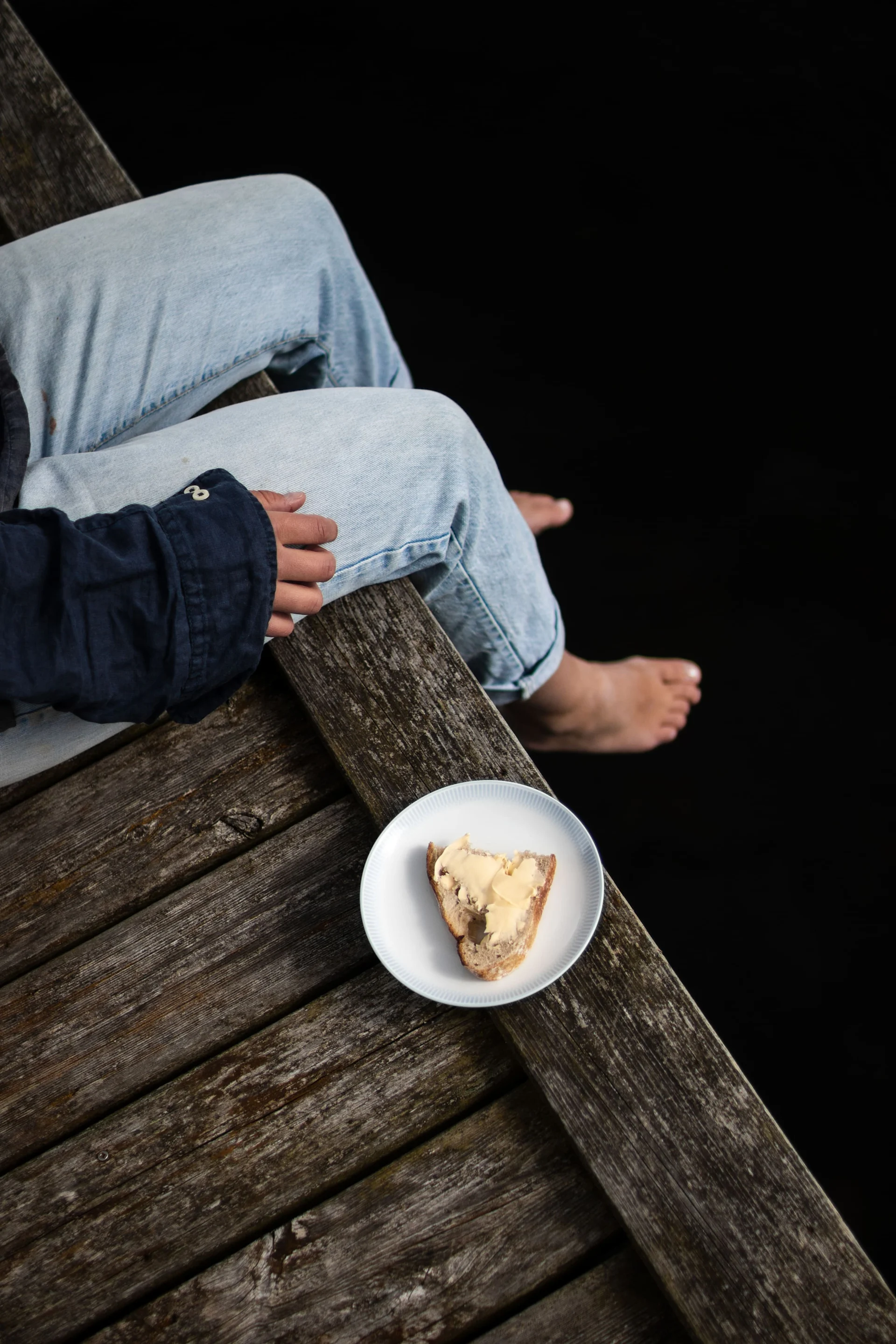 Persoon zit op een houten steiger met blote voeten naast een bord brood met boter op een donkere achtergrond.
