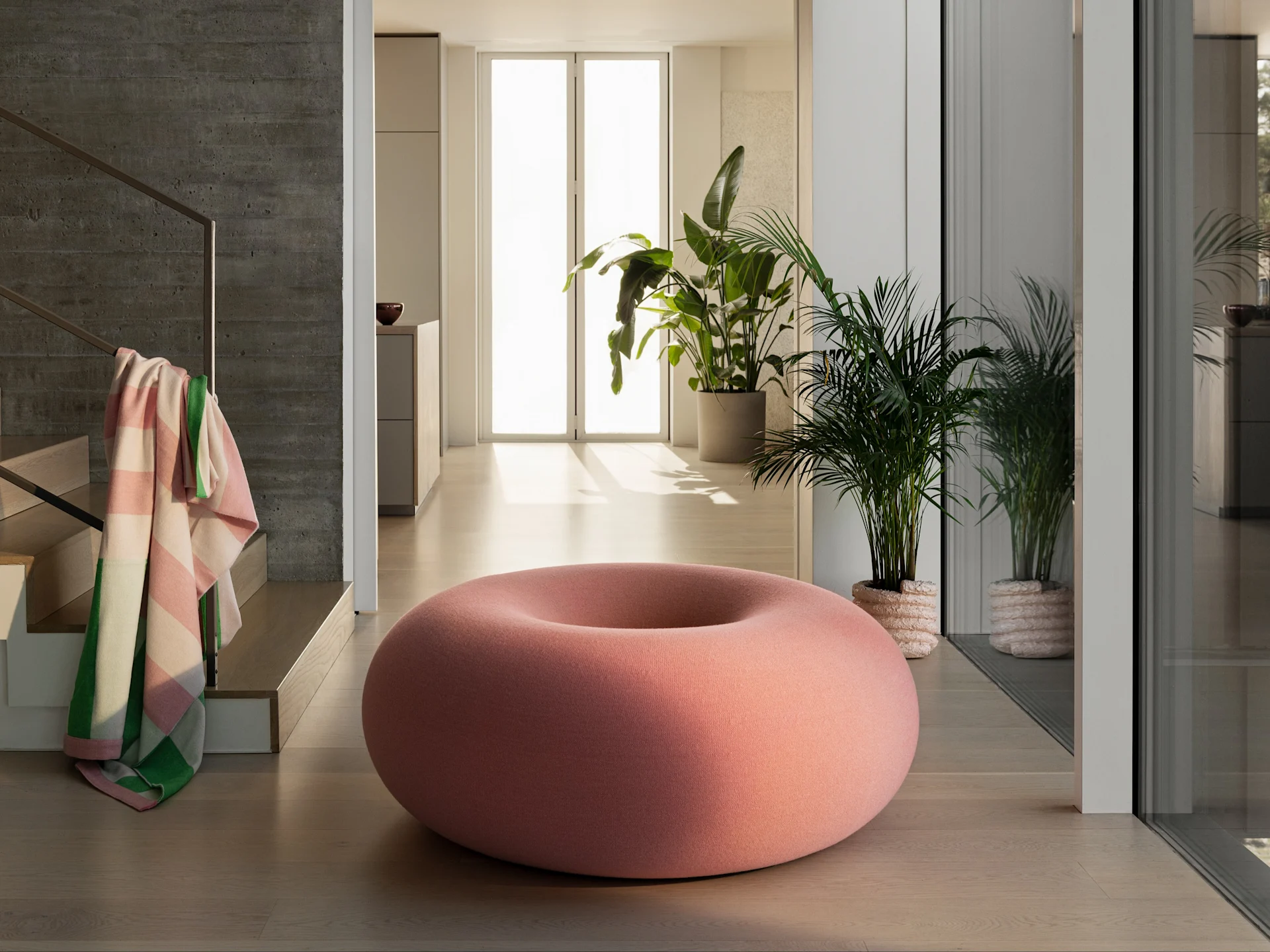 Een grote, donutvormige roze poef op een lichte houten vloer in een moderne kamer met groene planten en een betonnen muur.