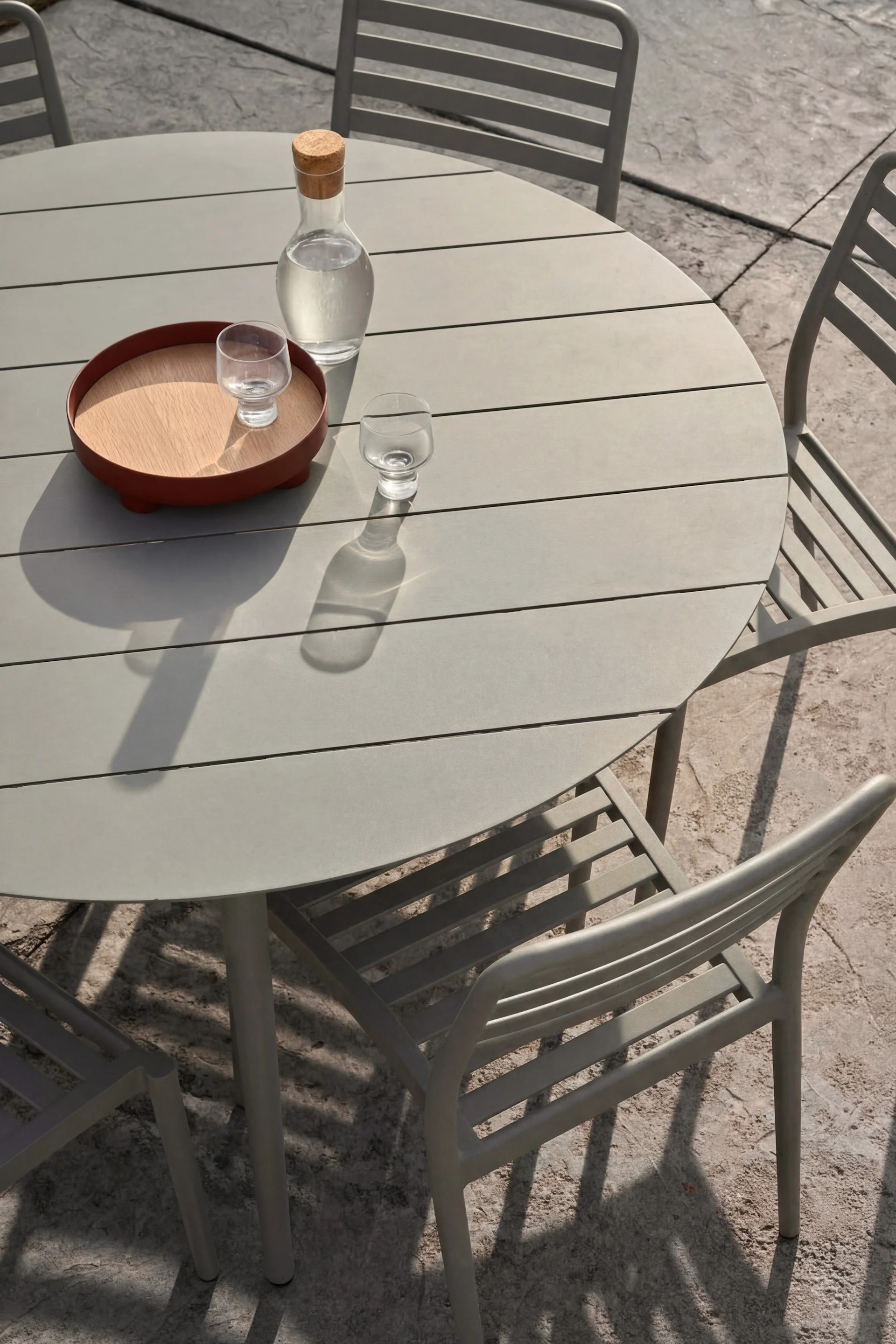 Ronde grijze buitentafel, drie stoelen. Glazen karaf water, twee glazen, één op houten dienblad, op tafel.