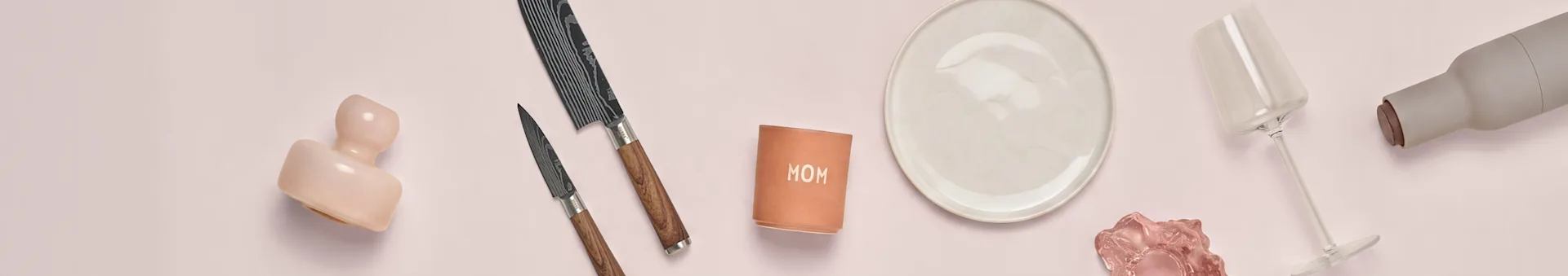 Roze keramiek, twee messen, 'MOM' beker, wit bord, roze glas, wijnglas, grijze fles op roze ondergrond.