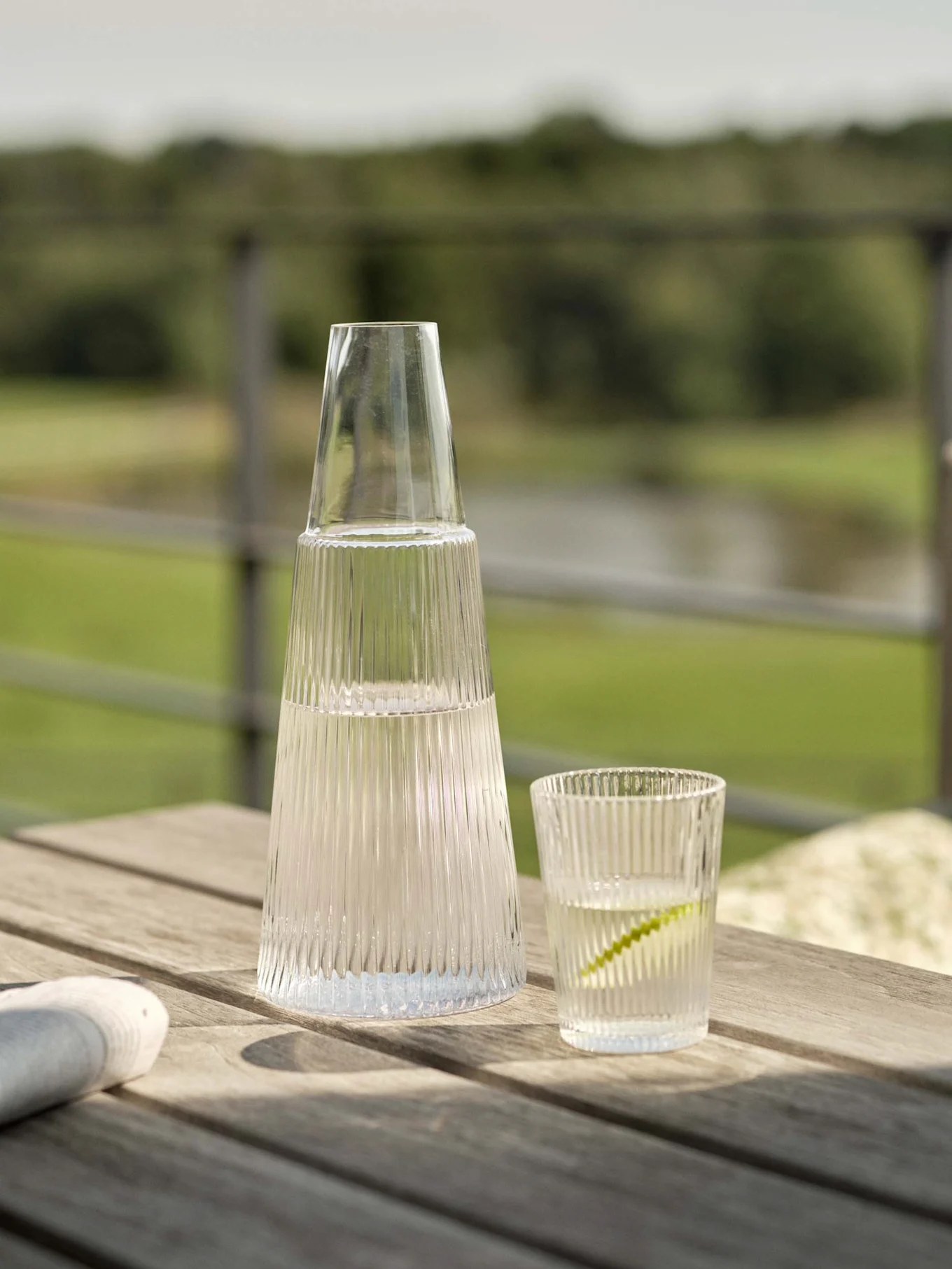Heldere geribbelde glazen karaf en bijpassend glas met water en limoen op een houten buitentafel.