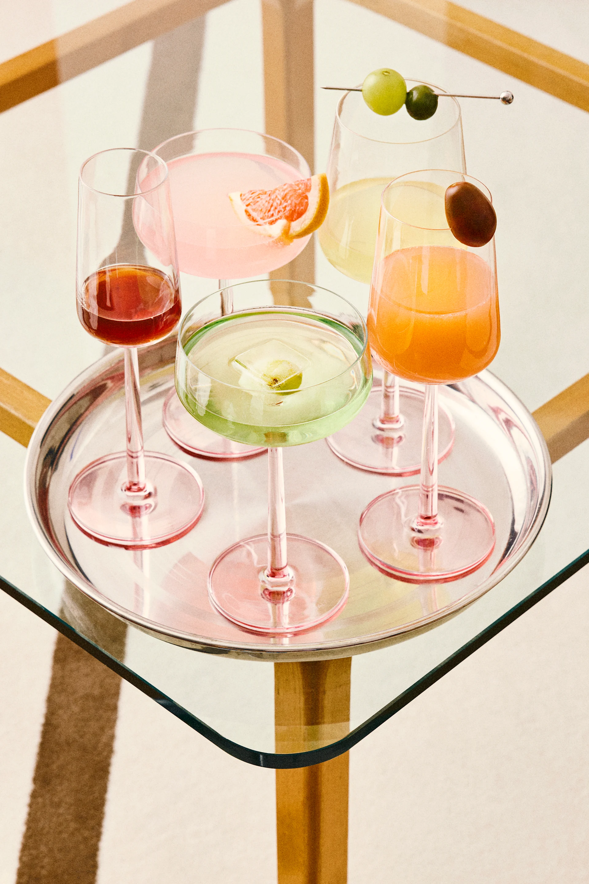 Vijf kleurrijke cocktails op een zilveren dienblad, waaronder roze, groene, oranje en rode drankjes met diverse garnituren, op een glazen tafel.