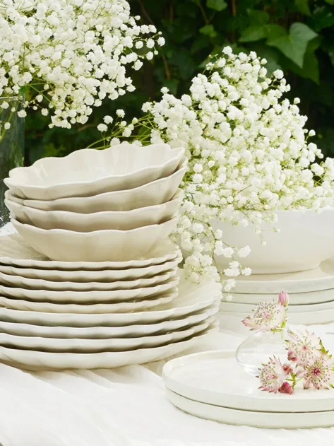 Gestapeld crèmekleurig keramisch servies met golvende randen, omringd door wit gipskruid en roze bloemen.