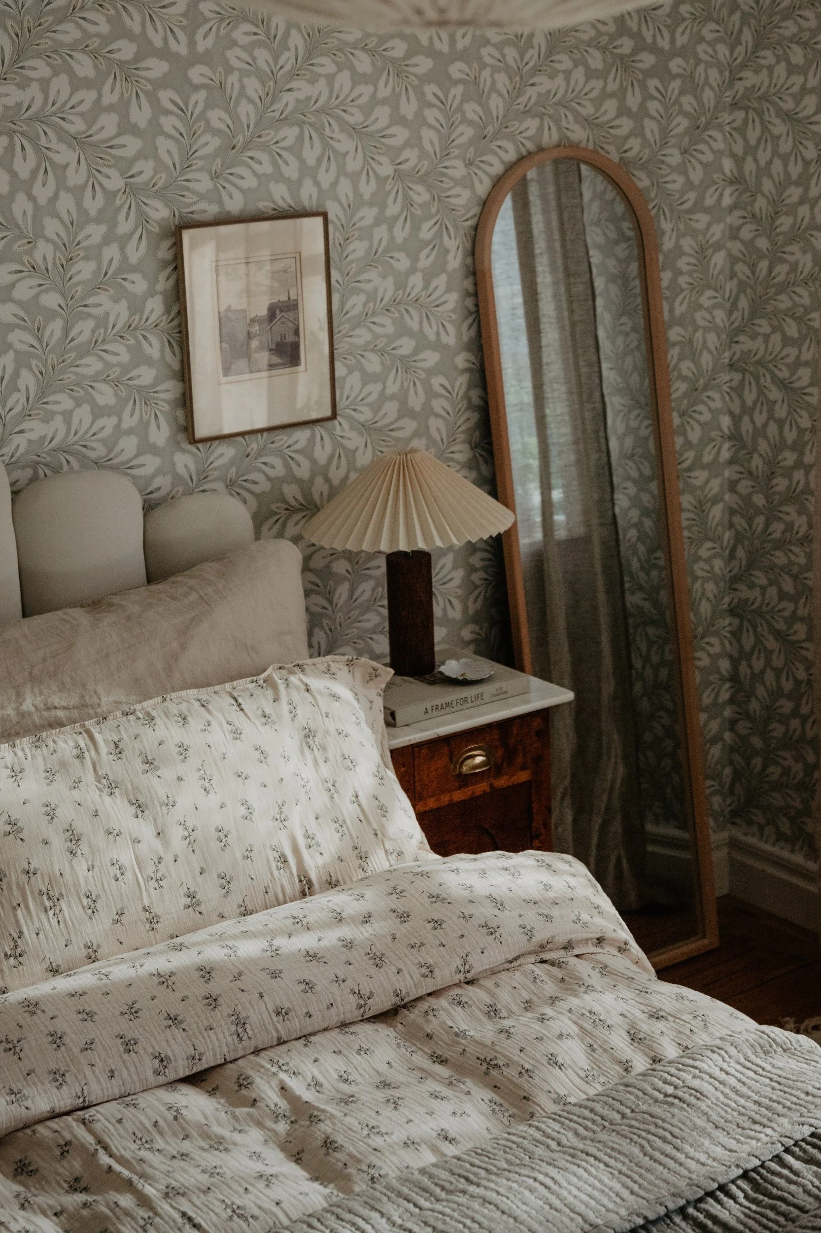 Een slaapkamer met crèmekleurig beddengoed met patroon, een geplooide lamp op een houten nachtkastje en een hoge spiegel, tegen bladbehang.