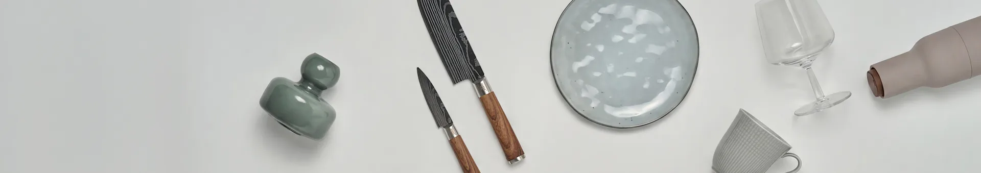 Diverse keuken- en eetartikelen, waaronder een groene vaas, twee messen, een gespikkeld bord, een wijnglas, een beige fles en een gemarmerde mok.