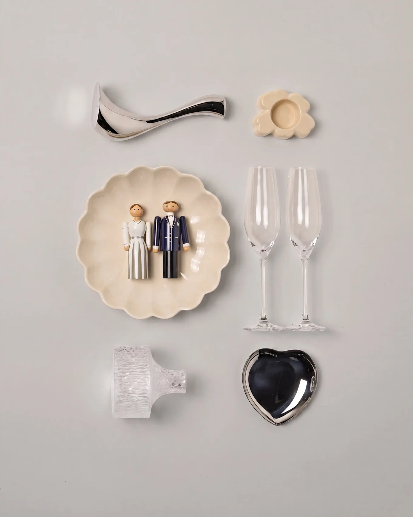 Huwelijks-flat lay met houten bruidspaarfiguren op een gegolfd bord, twee champagneglazen, een zilveren hartvormige schaal en diverse decoraties.