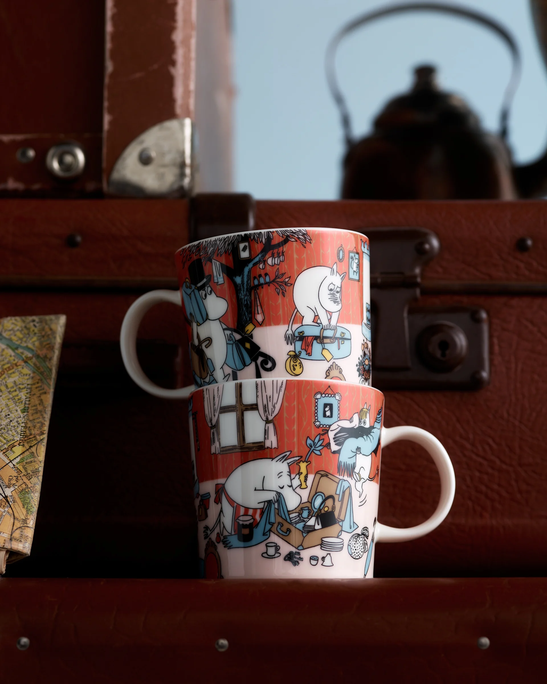 Twee Moomin-mokken met oranjerode karakterillustraties, gestapeld voor bruine koffers en een vage kaart.