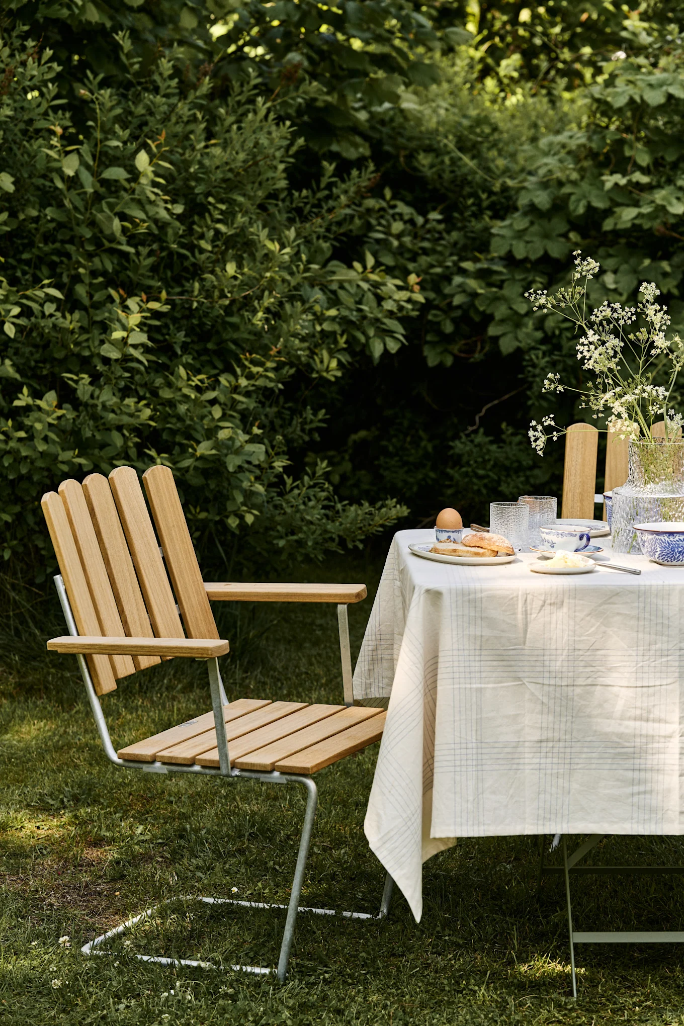 Een ontbijtscène buiten met een stoel van lichte houten latten en metaal naast een tafel gedekt met een geruite tafelkleed, servies en een vaas met witte bloemen.