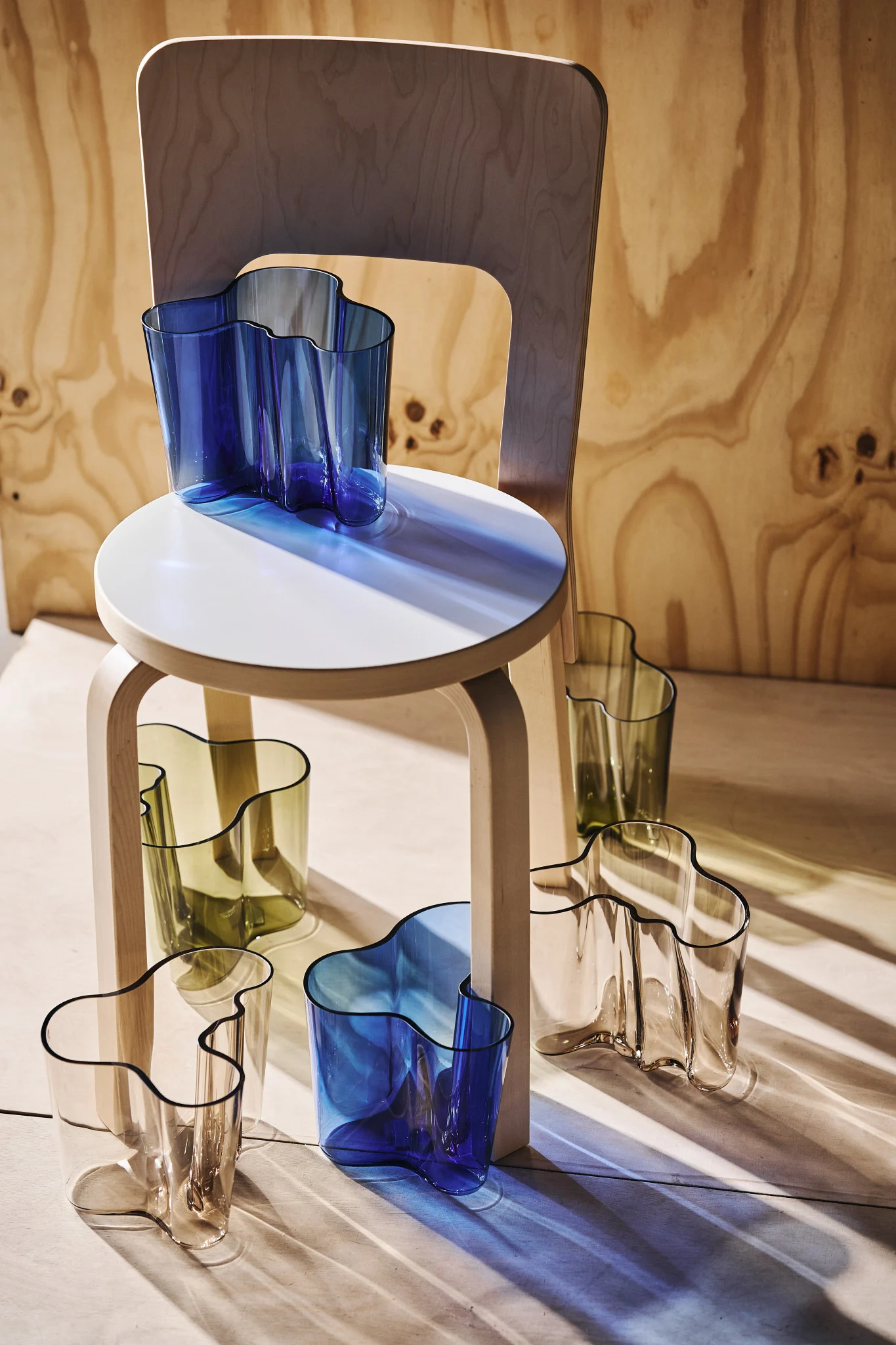 Meerdere golvende glazen vazen in blauw, olijfgroen en helder, gerangschikt op en rond een lichte houten kruk met een houten stoel op de achtergrond. Zonlicht werpt gekleurde schaduwen.