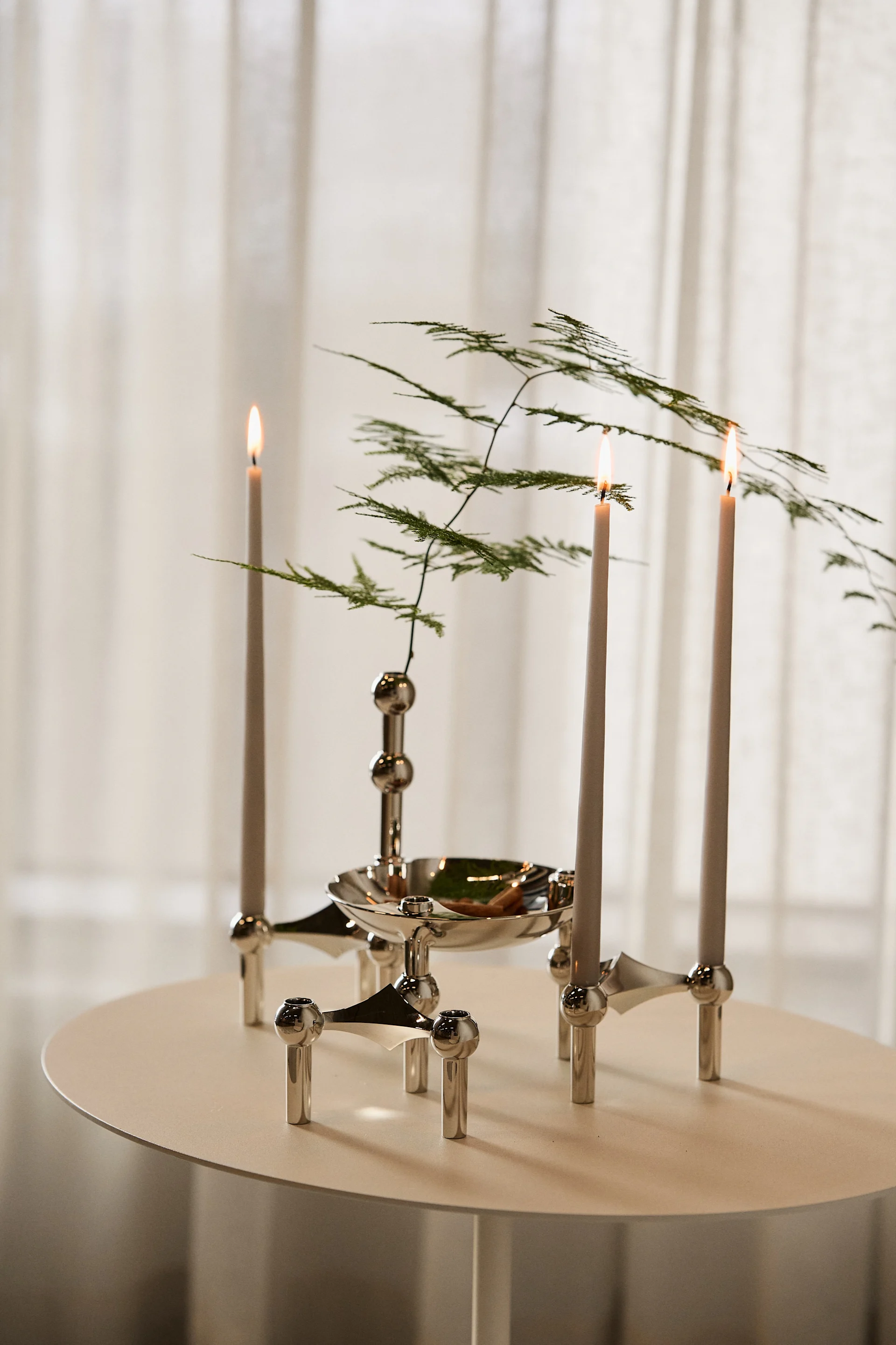 Drie brandende kaarsen in een modulaire zilveren kandelaar met groen blad, op een lichte ronde tafel.