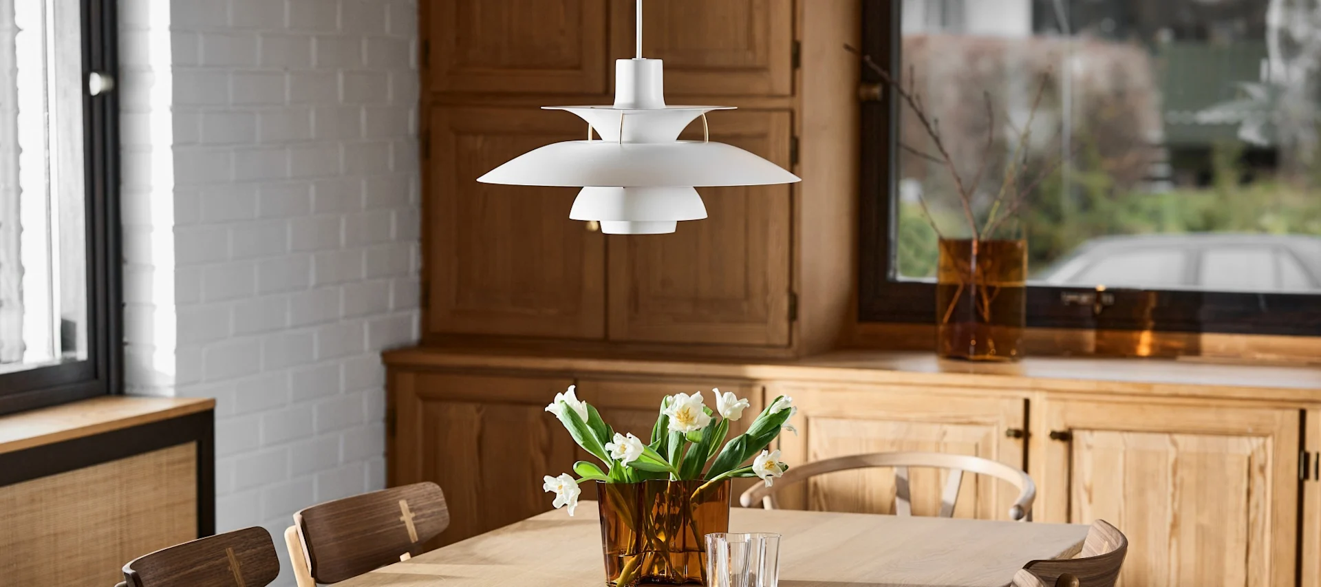 Een witte, meerlaagse hanglamp hangt boven een houten tafel met een vaas witte tulpen, in een kamer met houten kasten.