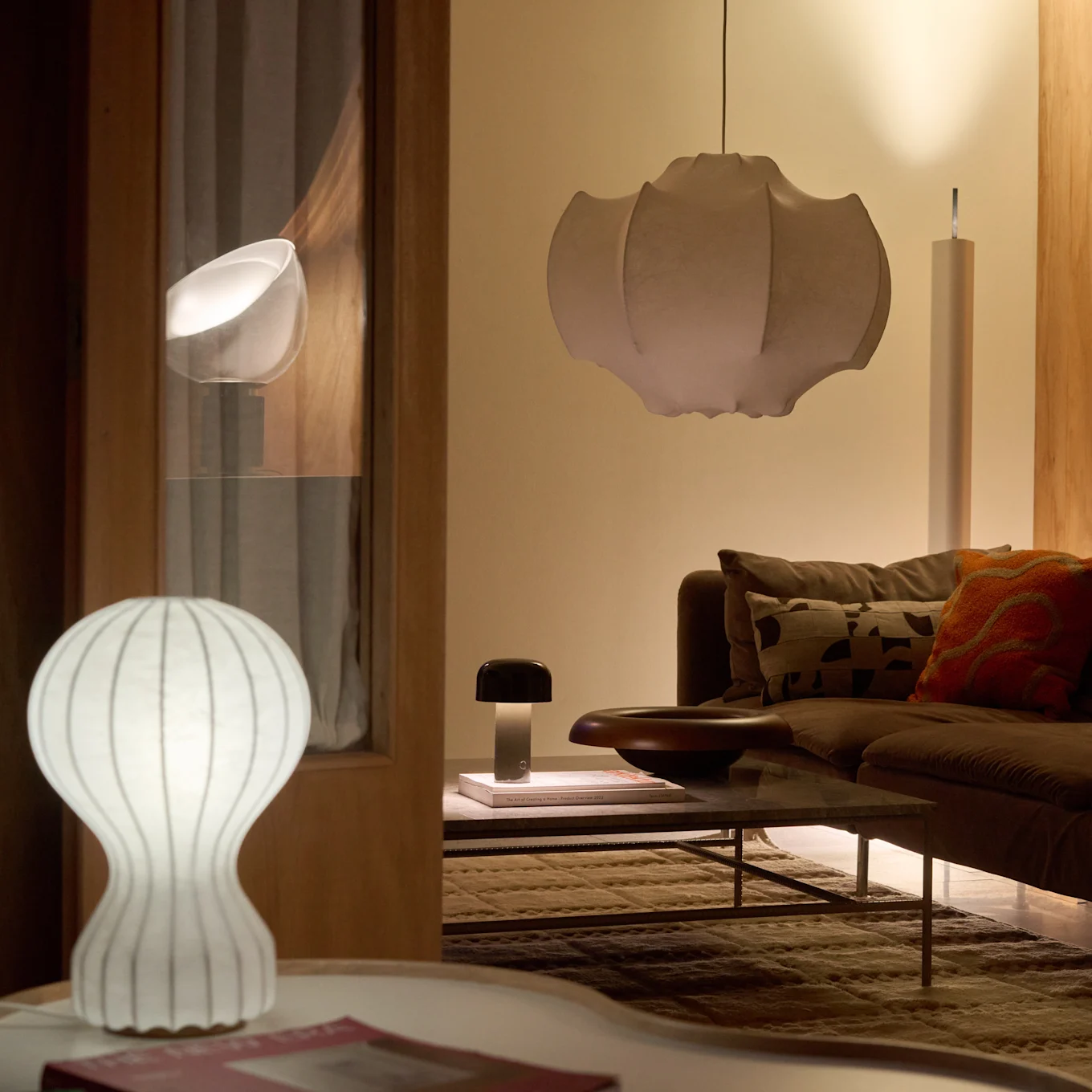Een woonkamer met een bruine bank en salontafel, verlicht door een witte geribbelde tafellamp, een donkere paddenstoellamp en een grote geplooide hanglamp, wat een warme gloed creëert.