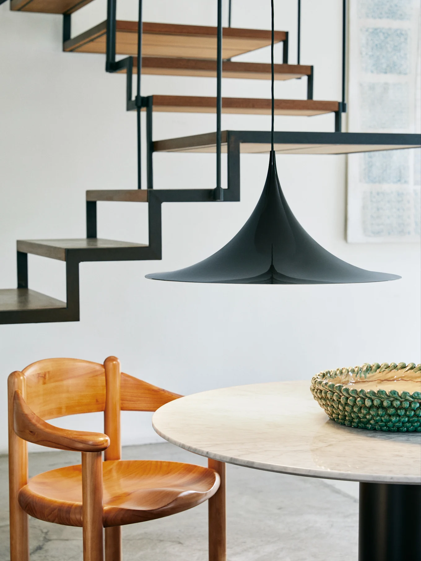 Een zwarte kegelvormige hanglamp hangt boven een ronde marmeren tafel en een houten stoel, met een moderne trap op de achtergrond.