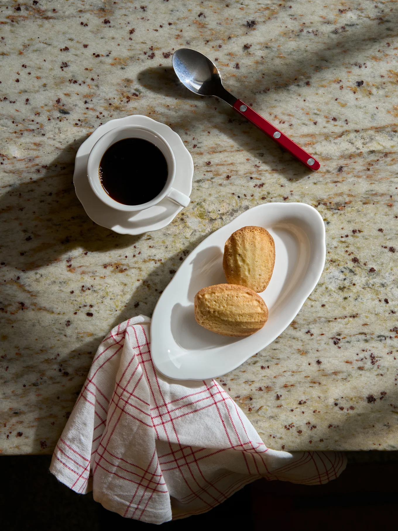 Zwarte koffie in witte kop, twee madeleines op wit bord, lepel en gestreepte servet op marmeren blad.